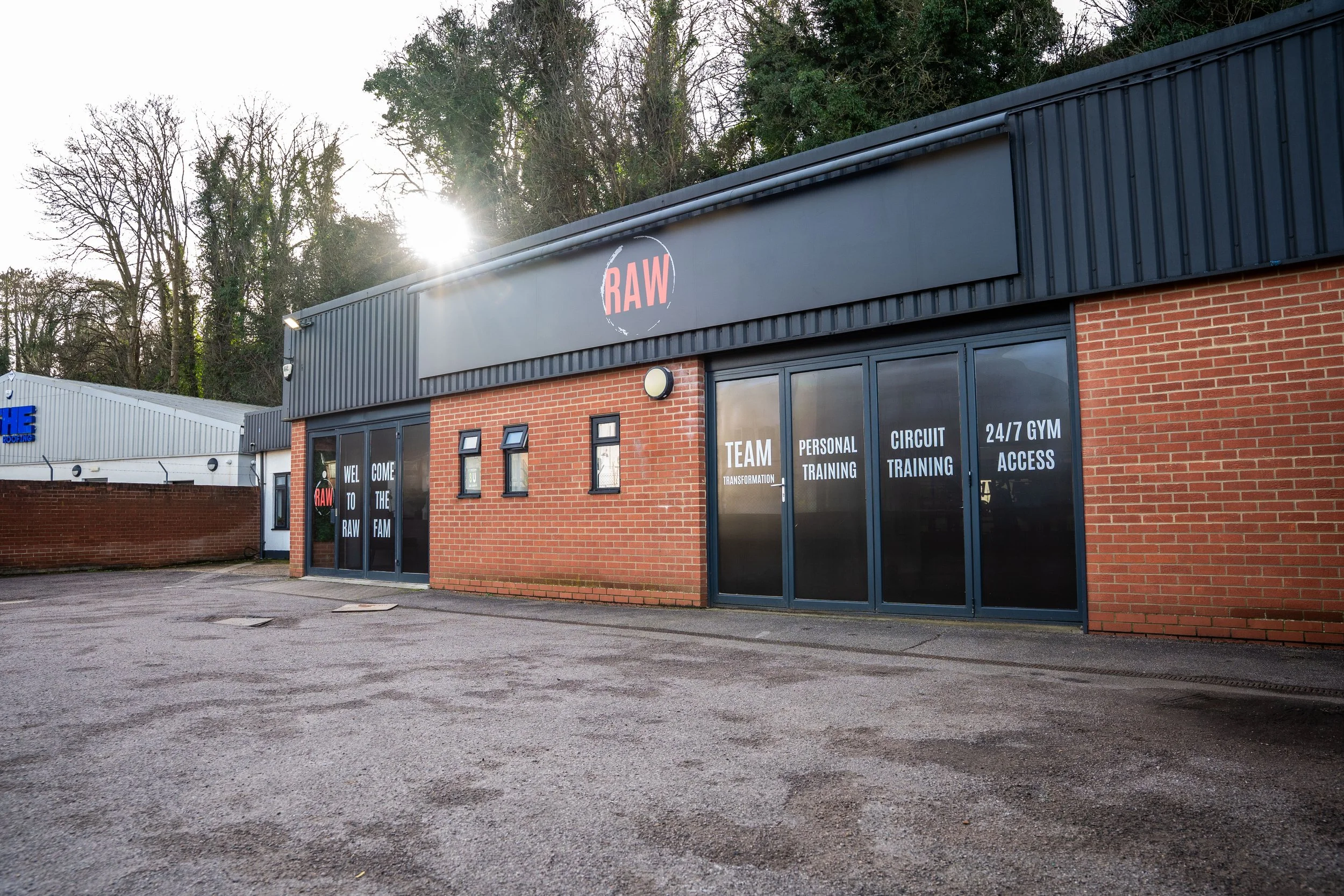 Exterior of a gym with black and brick facade, large glass doors with motivational signs, and a sign that says RAW, with trees in the background.