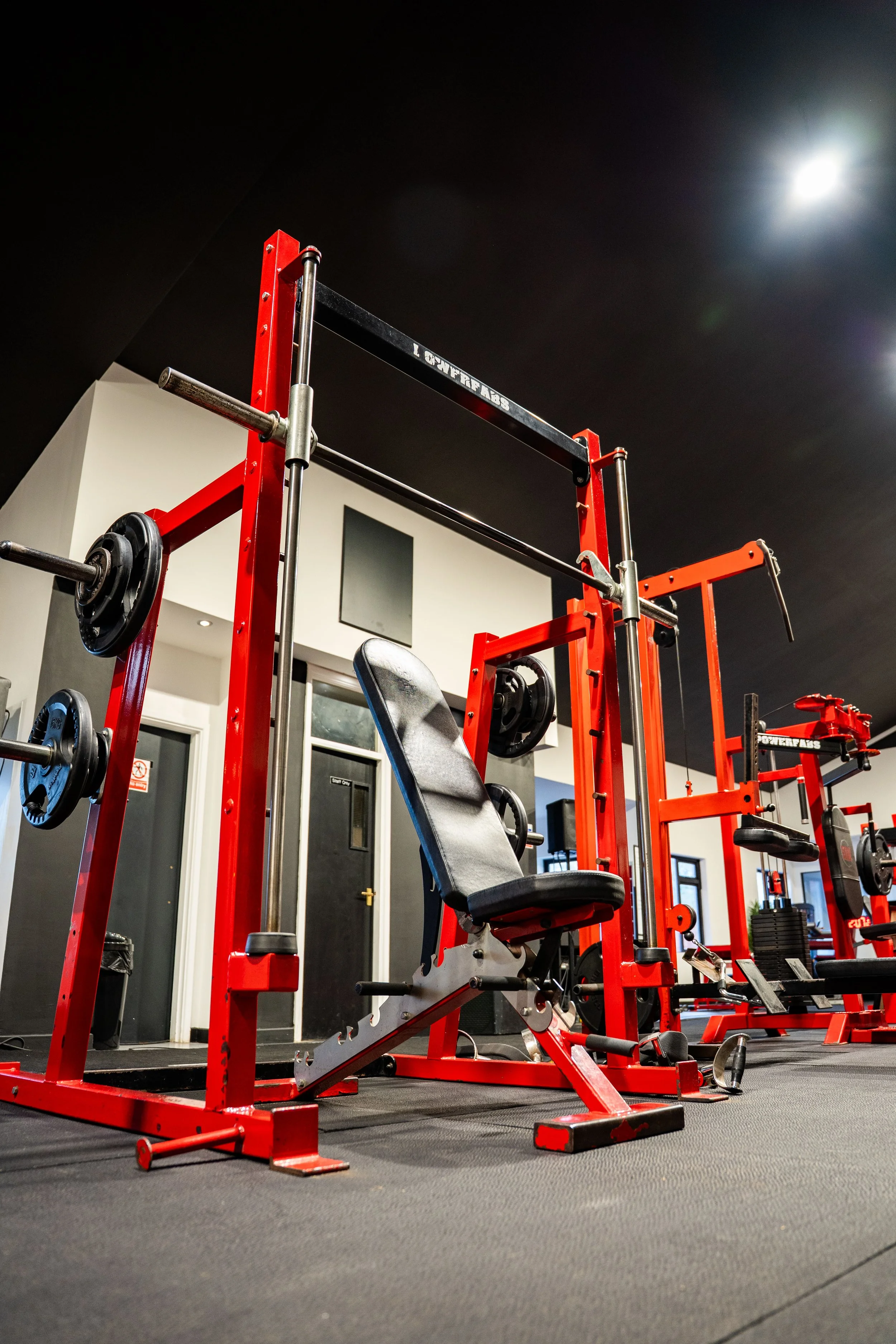 Empty gym with red weightlifting equipment on a black rubber floor under a black ceiling, illuminated by spotlights.