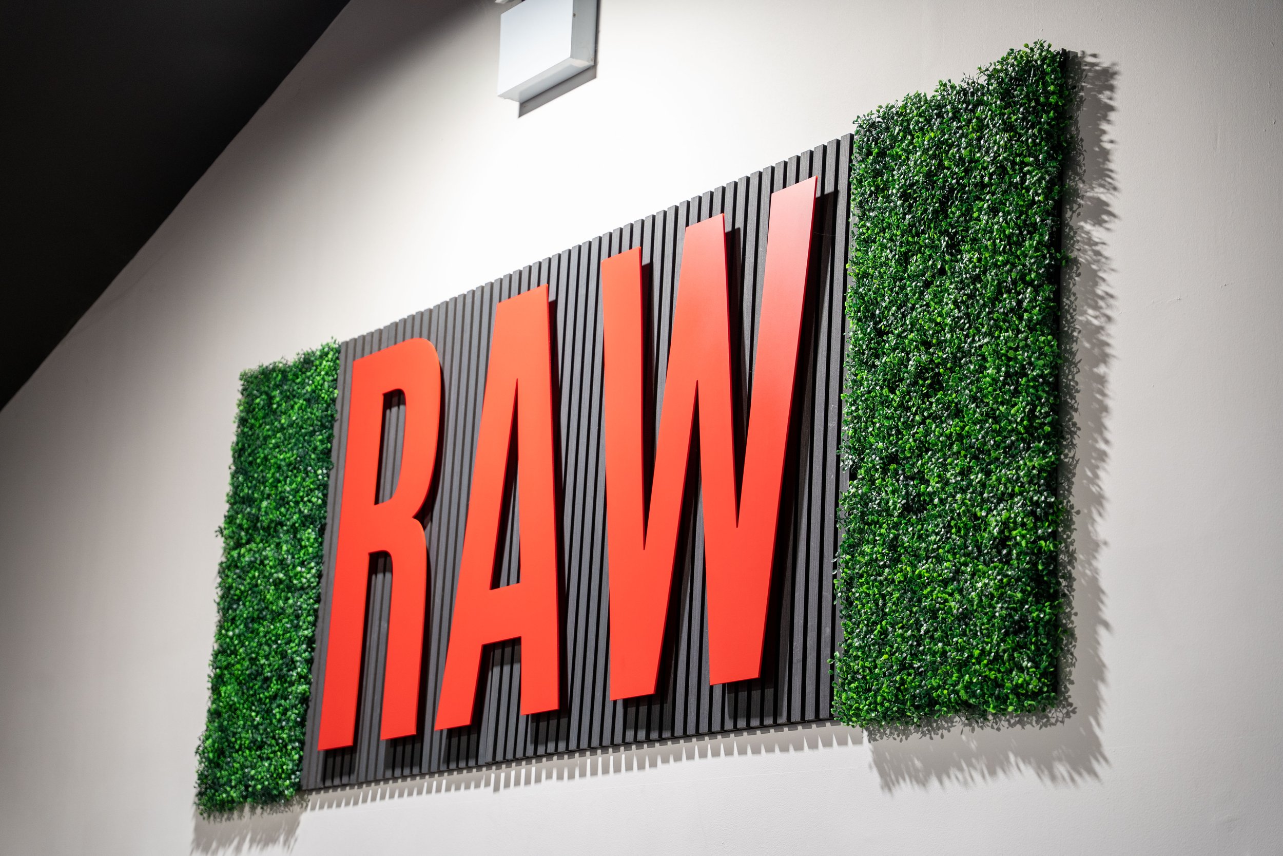 A sign with large red letters spelling 'RAW' on a black slatted background, flanked by green artificial foliage panels, mounted on a white wall.
