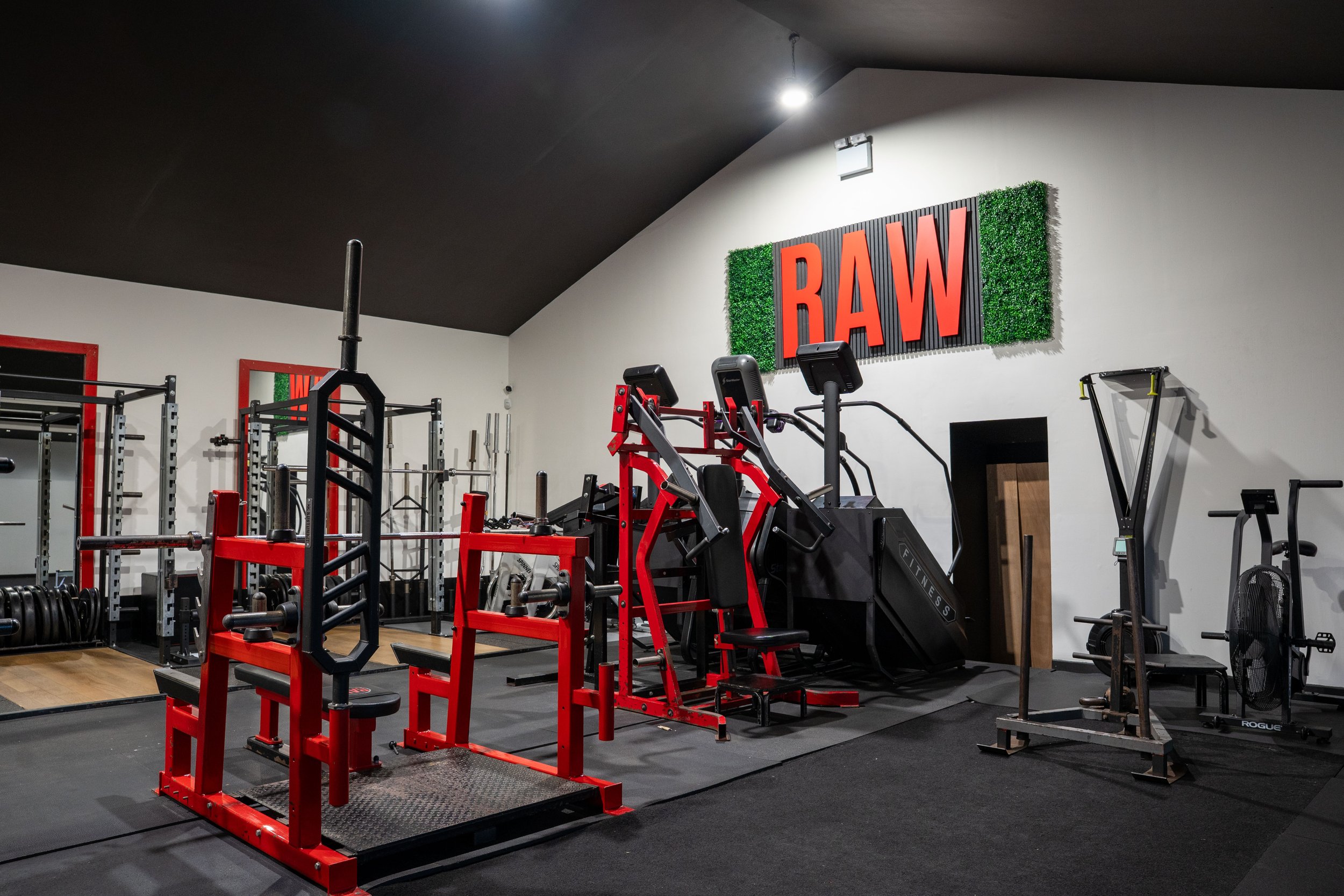 A gym with various exercise equipment including a leg press machine, weight racks, and a pitch-black wall with a large red RAW sign and green decorative border.