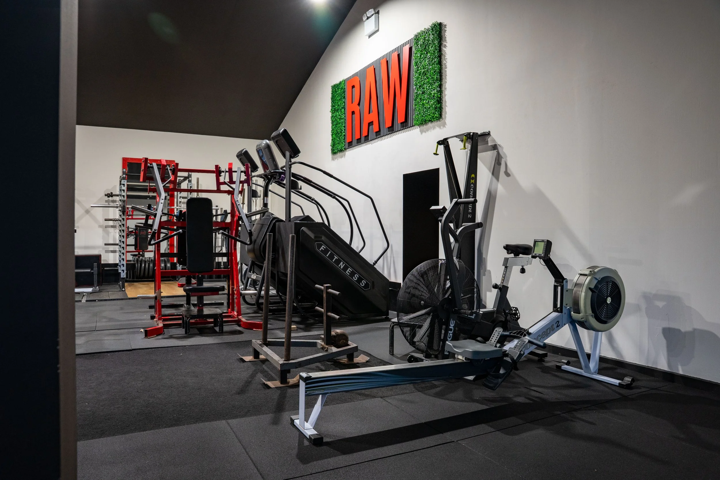 Gym with exercise equipment including a rowing machine, stationary bike, treadmill, and weight rack, with a sign that says 'RAW' on the wall.