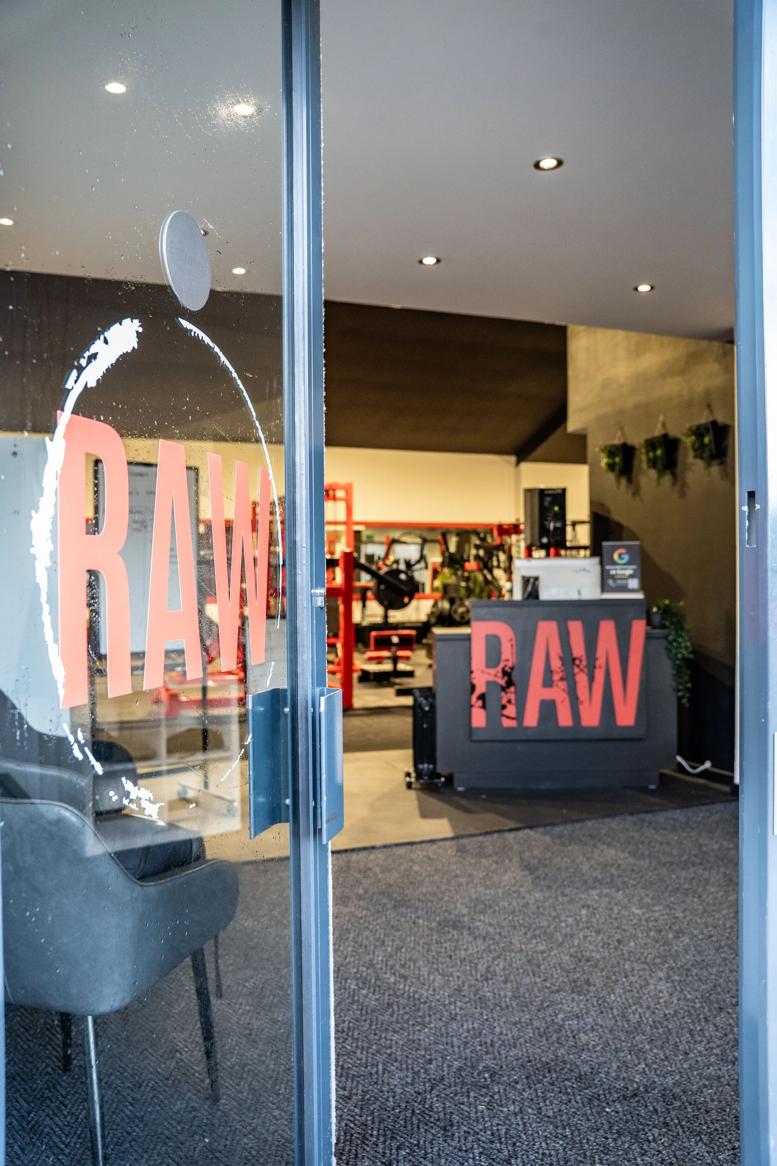 The interior of a fitness or wellness facility with a reception desk, workout equipment, and the word 'RAW' displayed on the glass door and the front of the desk.