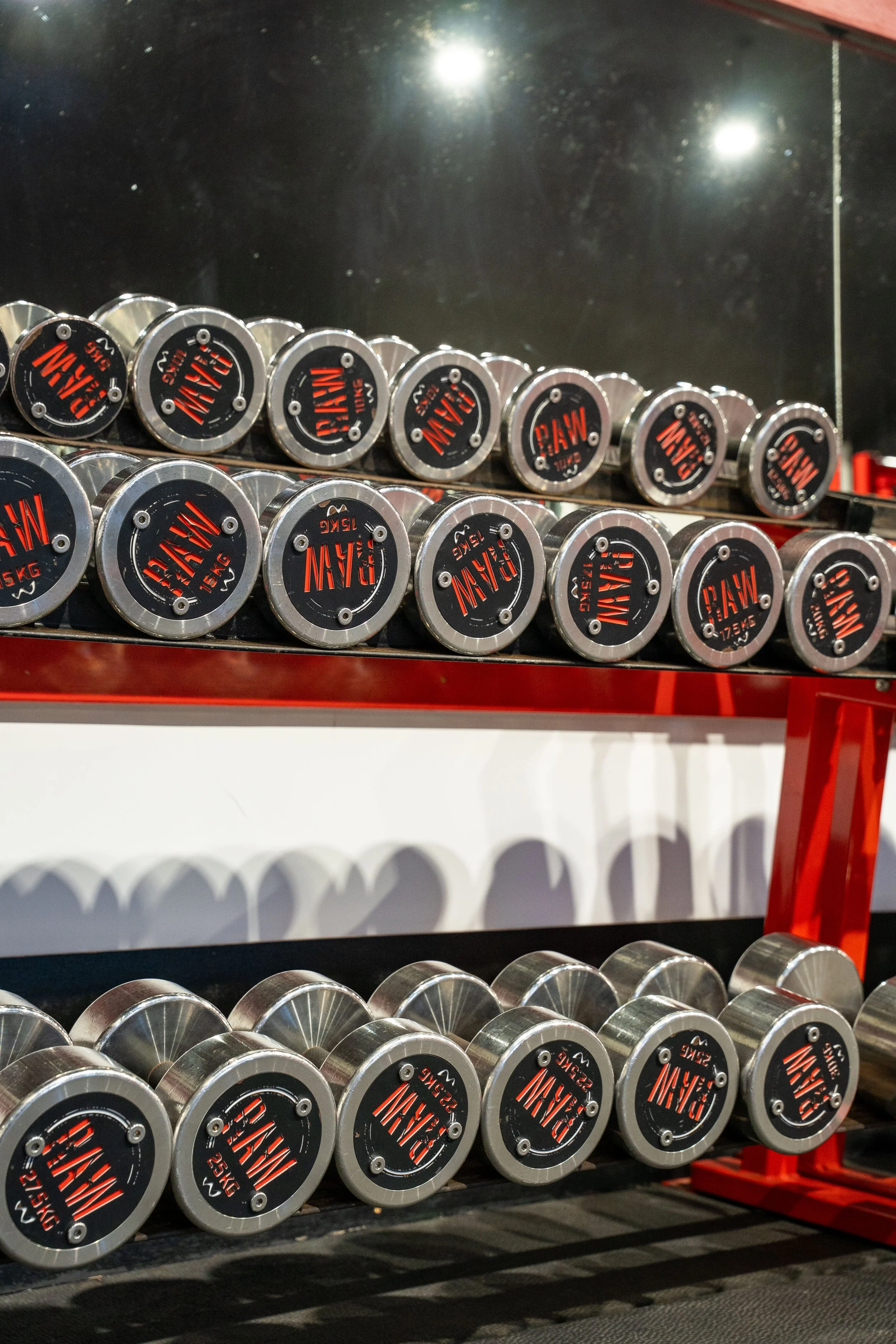 Rows of metal dumbbells with black and red weight labels on a red rack in a gym, with a mirror and reflection in the background.
