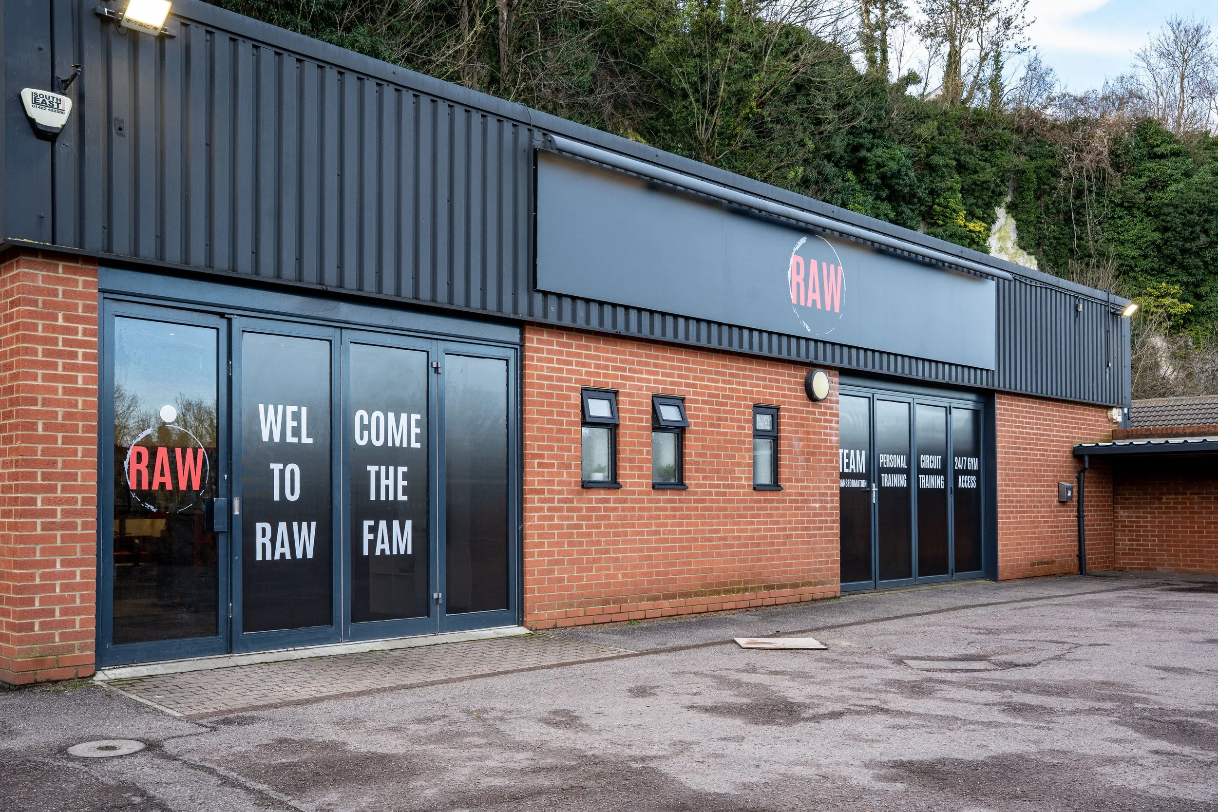 Exterior of a building with brick walls and blue doors, with signage indicating a fitness or training facility called RAW, featuring words like 'Welcome to the Raw', 'Team', 'Personal Training', 'Circuit Training', and '24/7 Gym Access'.