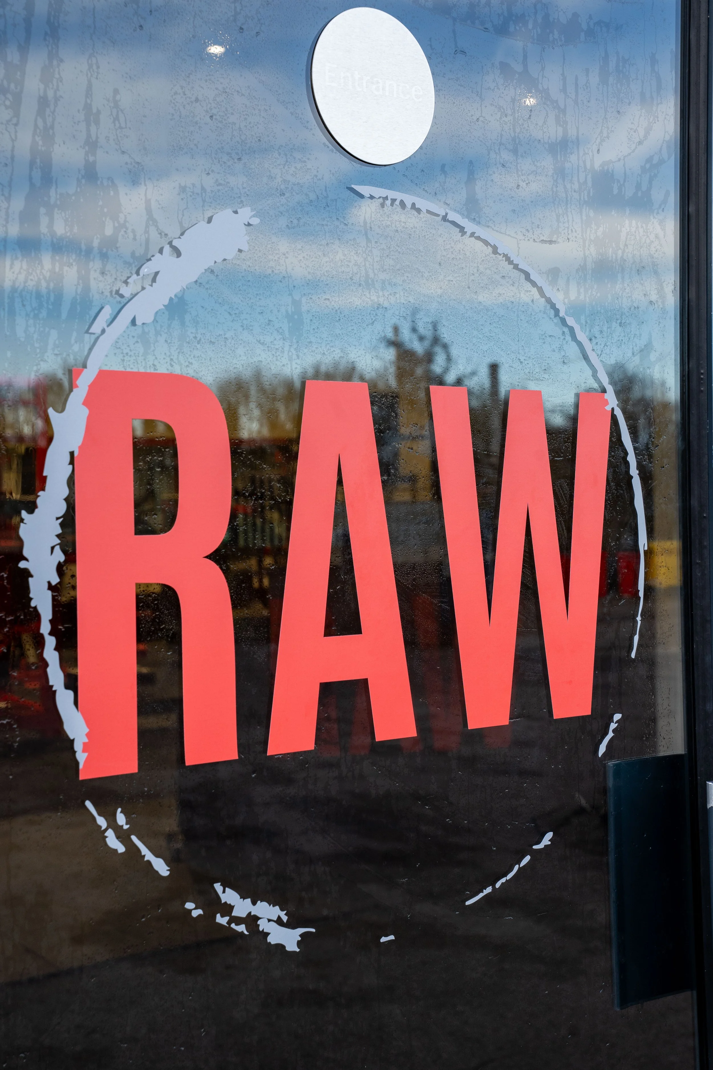 Store window with a red 'RAW' sign and a white circle above with the word 'Entrance' on it. The window is foggy with water streaks, showing trees and a cloudy sky in the background.
