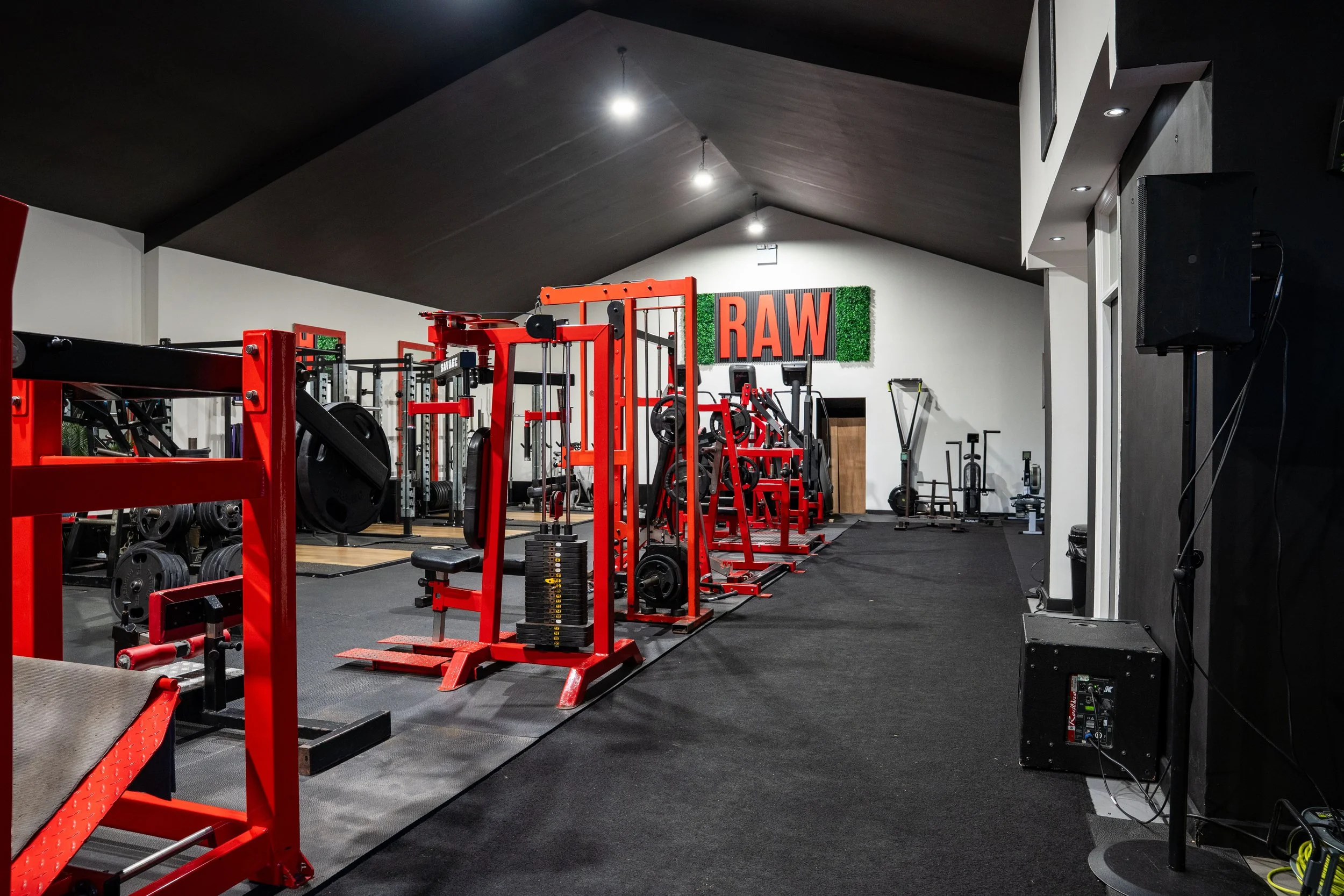 Interior view of a gym with various red weightlifting machines and free weights, and a sign on the wall that reads 'RAW' with green decorative background.