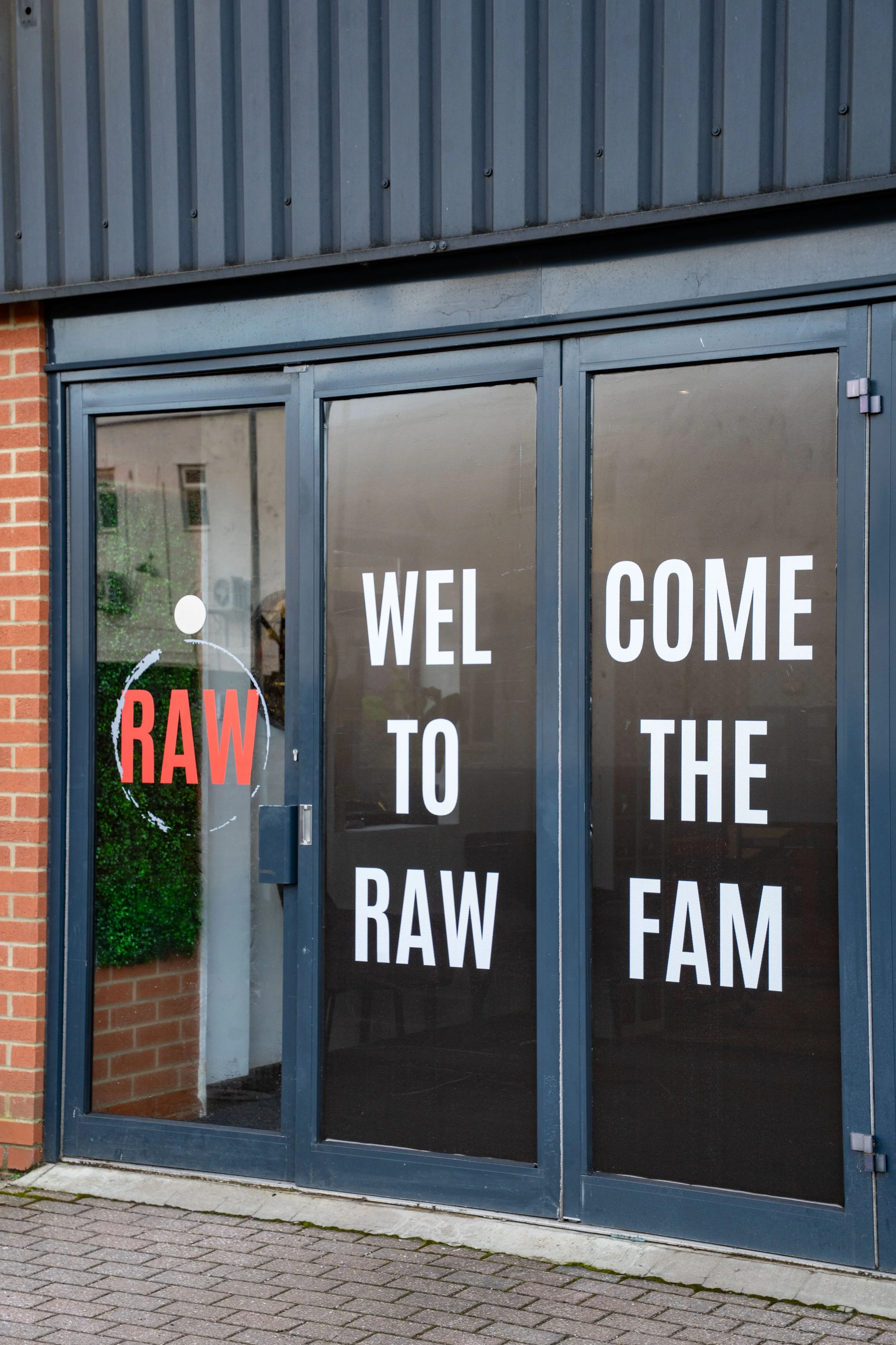 Storefront with glass doors displaying a sign that says "Welcome to the Raw Fam" and a logo with the word "RAW" in red and white.