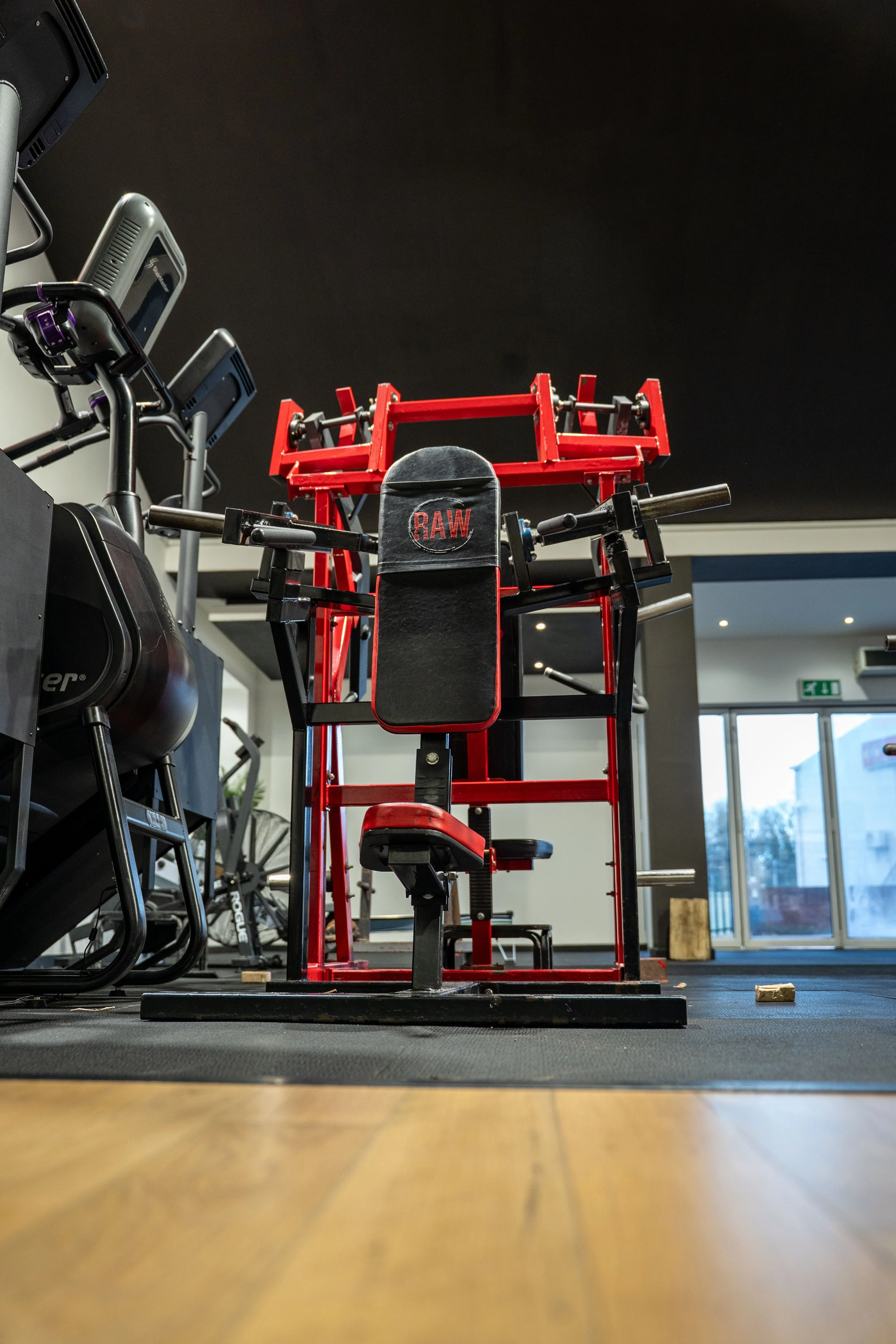 Gym equipment including a red weightlifting machine, stationary bikes on the left, and gym mats on the floor, with a view of large windows in the background.
