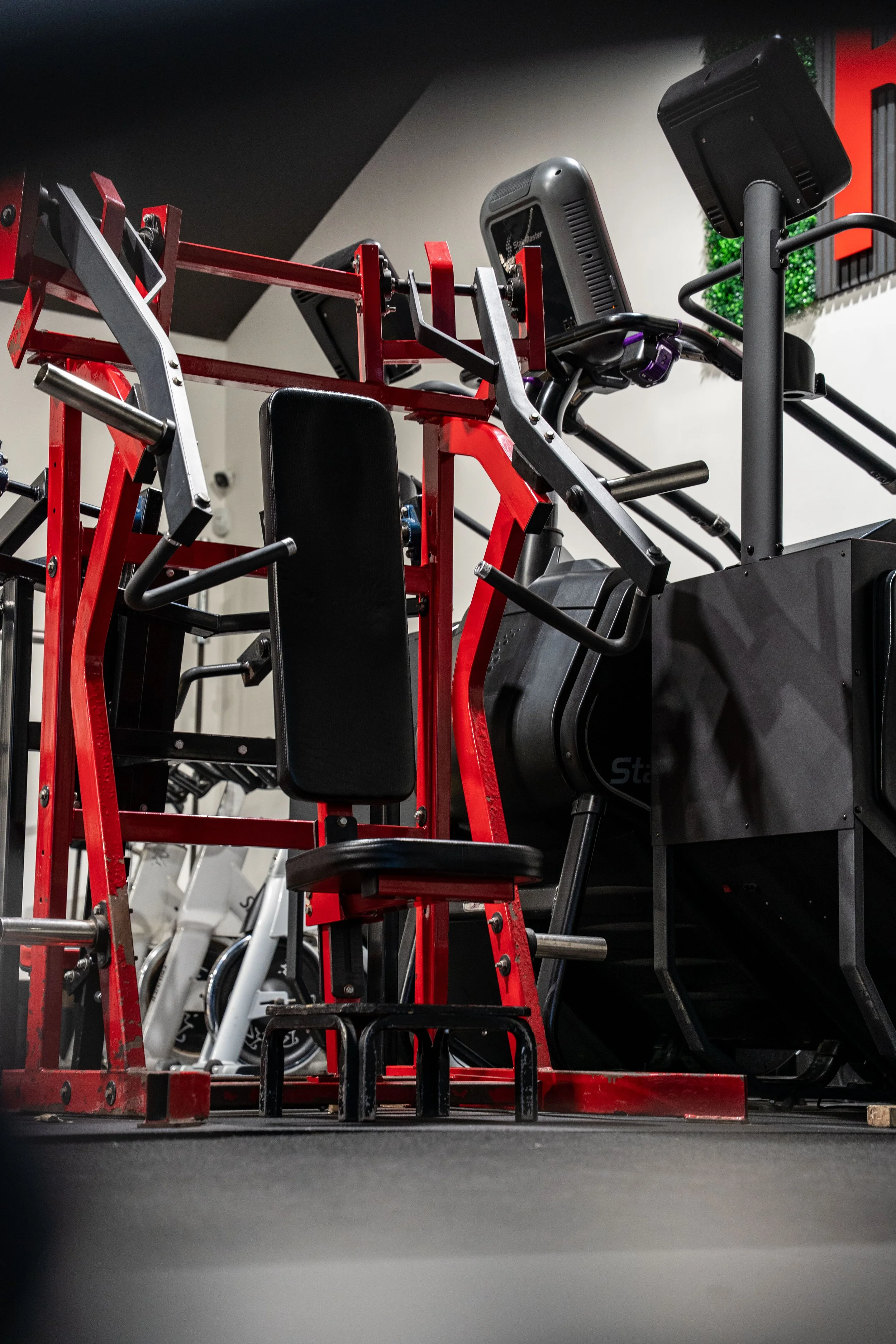 A fitness gym with various workout equipment, including a red and black strength training machine and an exercise bike, on a dark gym floor.