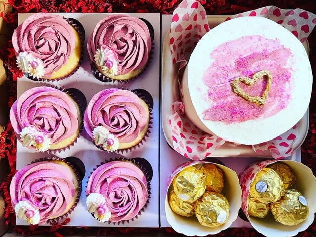 A gift box containing six pink frosted cupcakes with decorative flowers, a small cake with a pink and gold heart design, and several gold-wrapped chocolates.