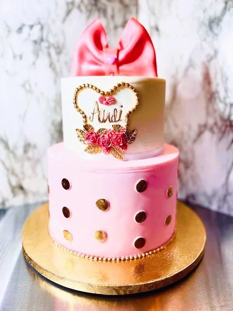 Two-tier pink and white cake with a large pink bow topper and gold polka dots. The top layer has a decorative gold frame with roses and a personalized name, resting on a golden cake board.