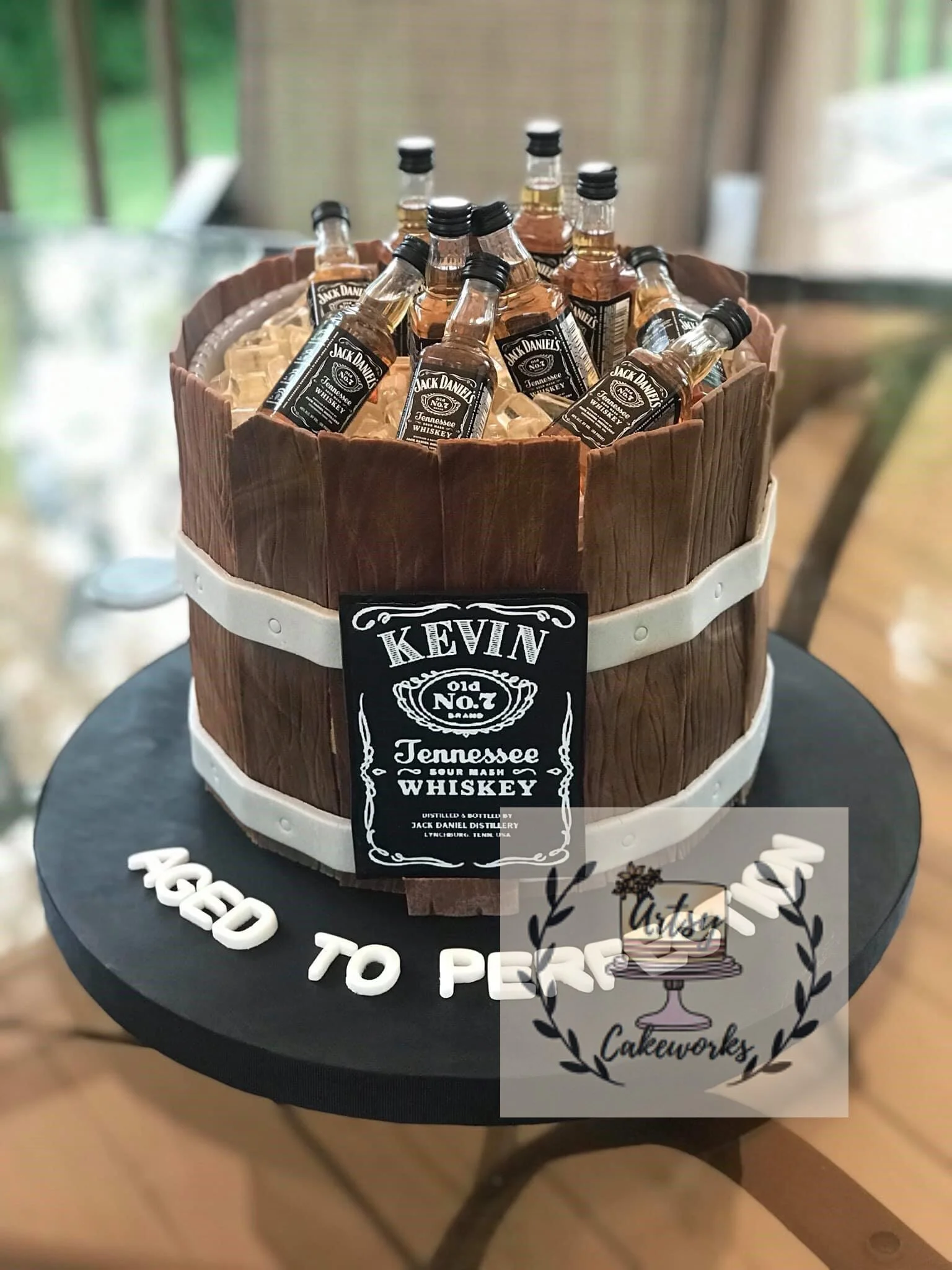 A cake designed to look like a wooden barrel filled with miniature bottles of Jack Daniel's Tennessee whiskey. The cake features a realistic wood texture and a white band around the middle, with a label that reads "Kevin" in the style of a whiskey la