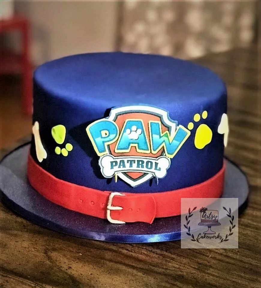 A cake decorated with a PAW Patrol theme, featuring a blue fondant cover, a red belt design around the base, and yellow paw prints. The PAW Patrol logo is prominently displayed on the side.