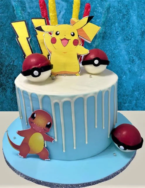 Blue and white drip cake featuring Pokémon-themed decorations with Pikachu and Charmander figures, Poké Balls, and lightning bolt designs.