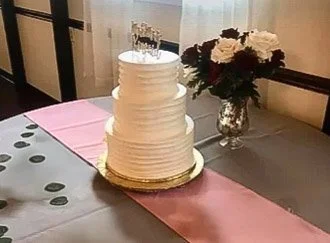 White tiered wedding cake on a table with a pink table runner and a flower arrangement in a glass vase.