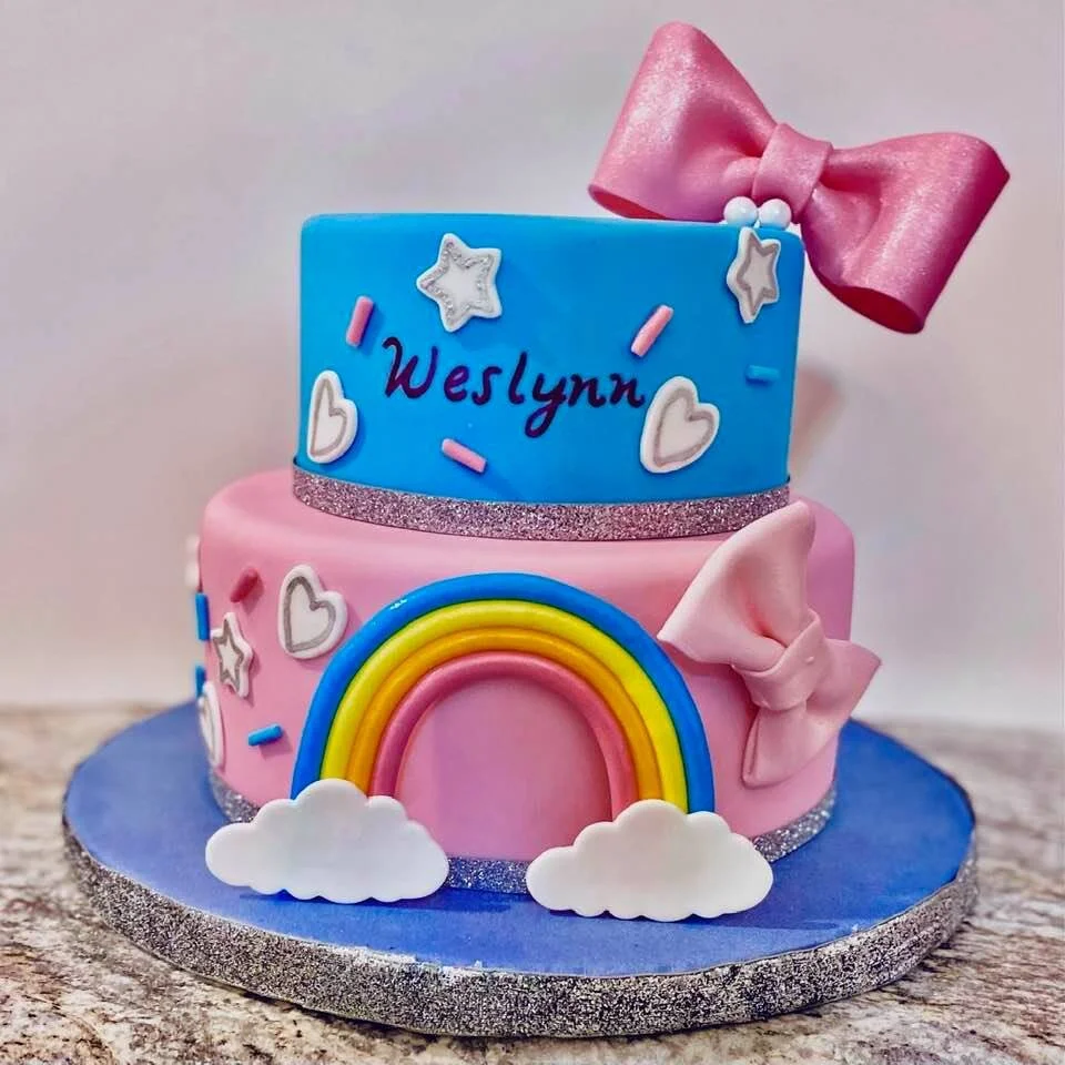 Two-tier cake with blue and pink fondant, decorated with bows, stars, hearts, and a rainbow. "Weslynn" is written on the top tier.