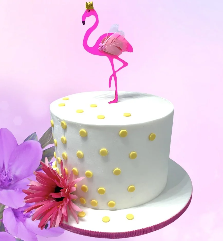 White cake with yellow polka dots, pink flamingo topper, and pink flowers on a pastel background.