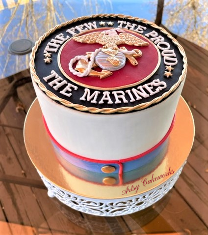 Cake with a Marine Corps emblem on top, featuring the phrase "The Few, The Proud, The Marines" and a red, white, and gold design, on a gold and white base.