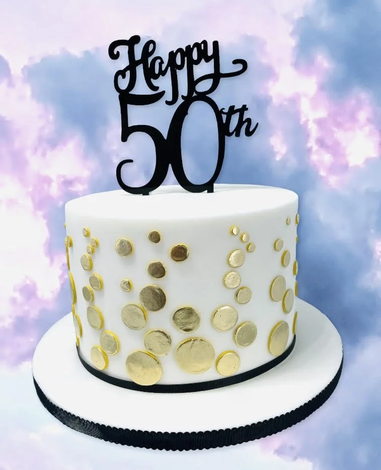 A white cake with gold circular decorations and a black ribbon trim topped with a "Happy 50th" topper against a cloudy background.