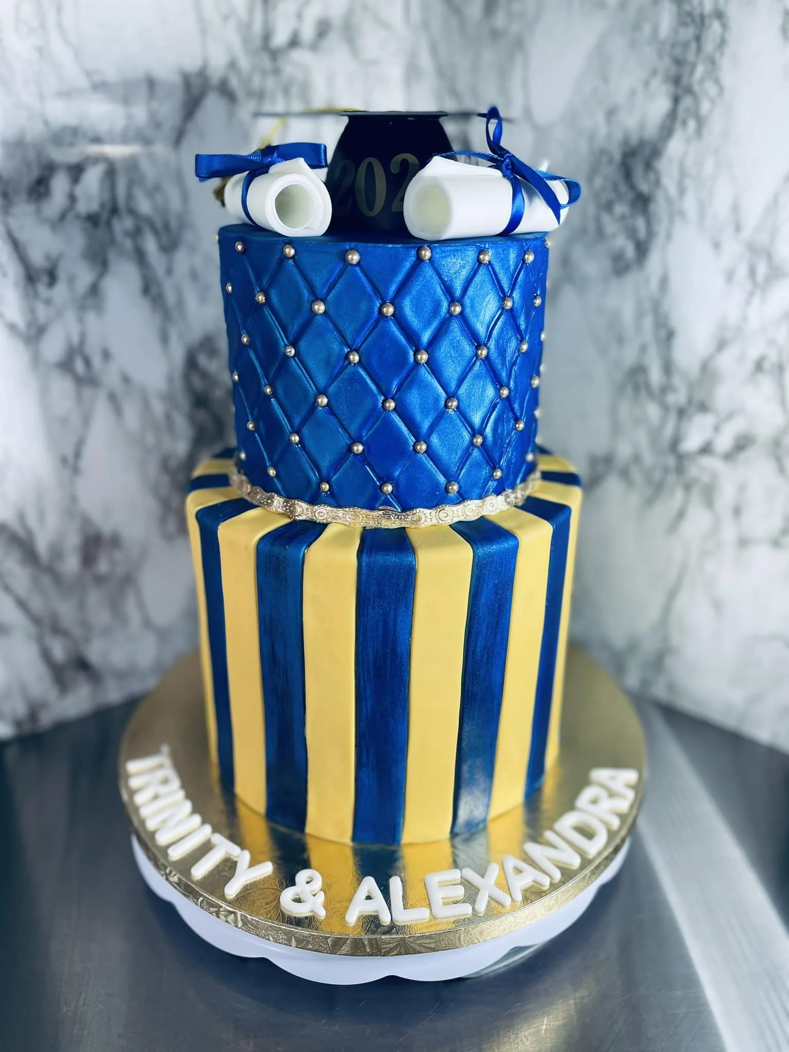 Three-tiered cake with striped yellow and blue bottom tier, quilted middle layer with silver beads, and top decoration of scrolls and a blue cap. Base reads "TRINITY & ALEXANDRA."