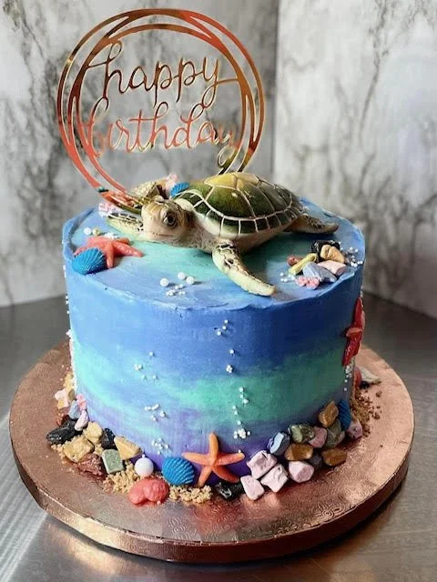 A marine-themed birthday cake with a blue and green water-like design, topped with a realistic turtle figurine, starfish, and colorful edible seashells and rocks. A "happy birthday" topper is on top.