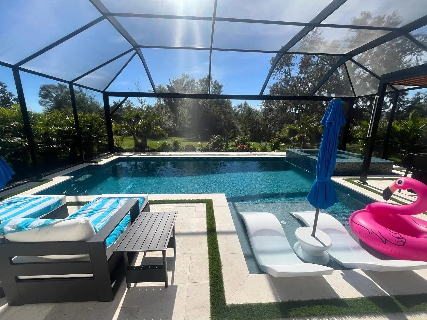 Backyard swimming pool enclosed by black mesh screening, with poolside lounge chairs with blue and white striped cushions, a pink flamingo pool float, blue umbrellas, and lush green trees and plants in the background.