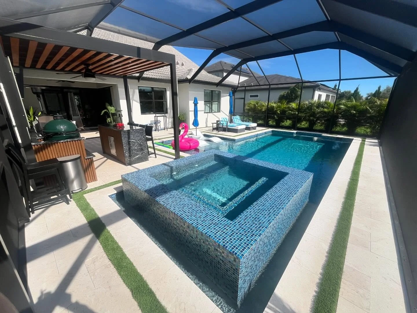 Luxury backyard with a screened-in pool area featuring a small hot tub, lounge chairs with blue umbrellas, a green rug, pink flamingo pool float, and an outdoor kitchen area with a green grill.