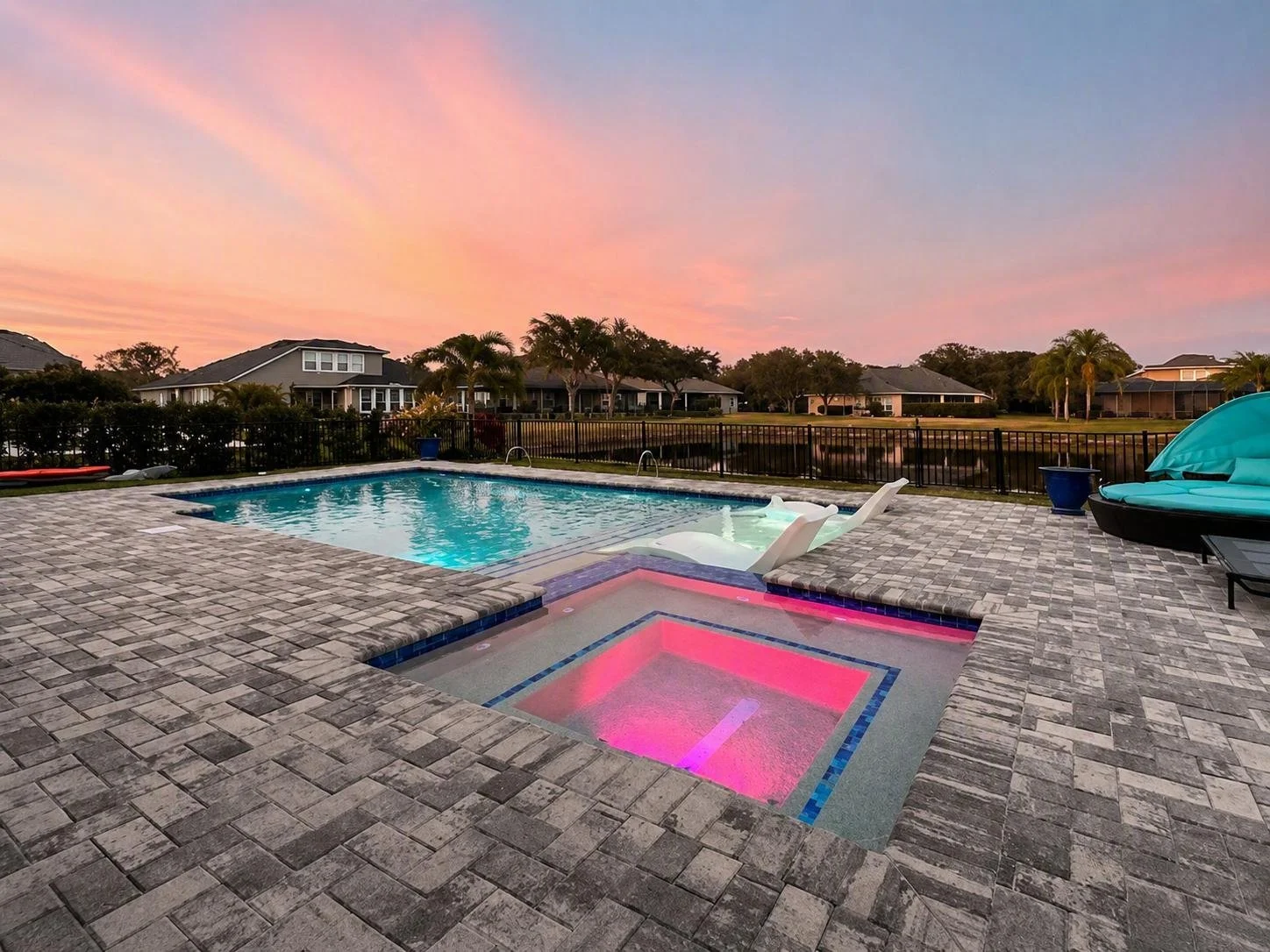A backyard pool area during sunset with a rectangular pool, a hot tub illuminated with pink and purple lighting, lounge chairs, and a blue canopy, surrounded by a brick patio and a black metal fence, with houses and palm trees in the background.