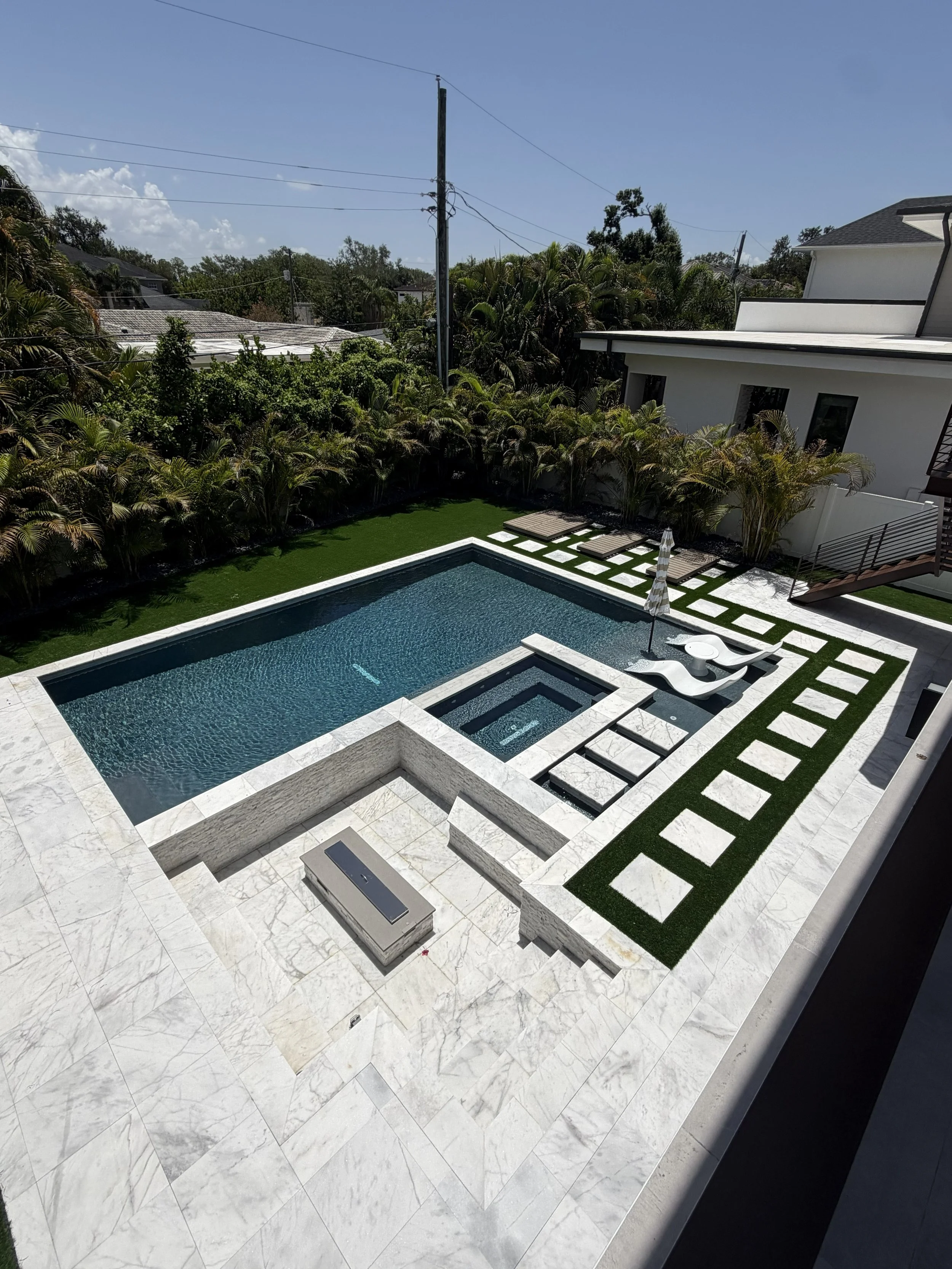 Modern backyard with swimming pool, lounge chairs, greenery, and neighboring houses under a clear blue sky.