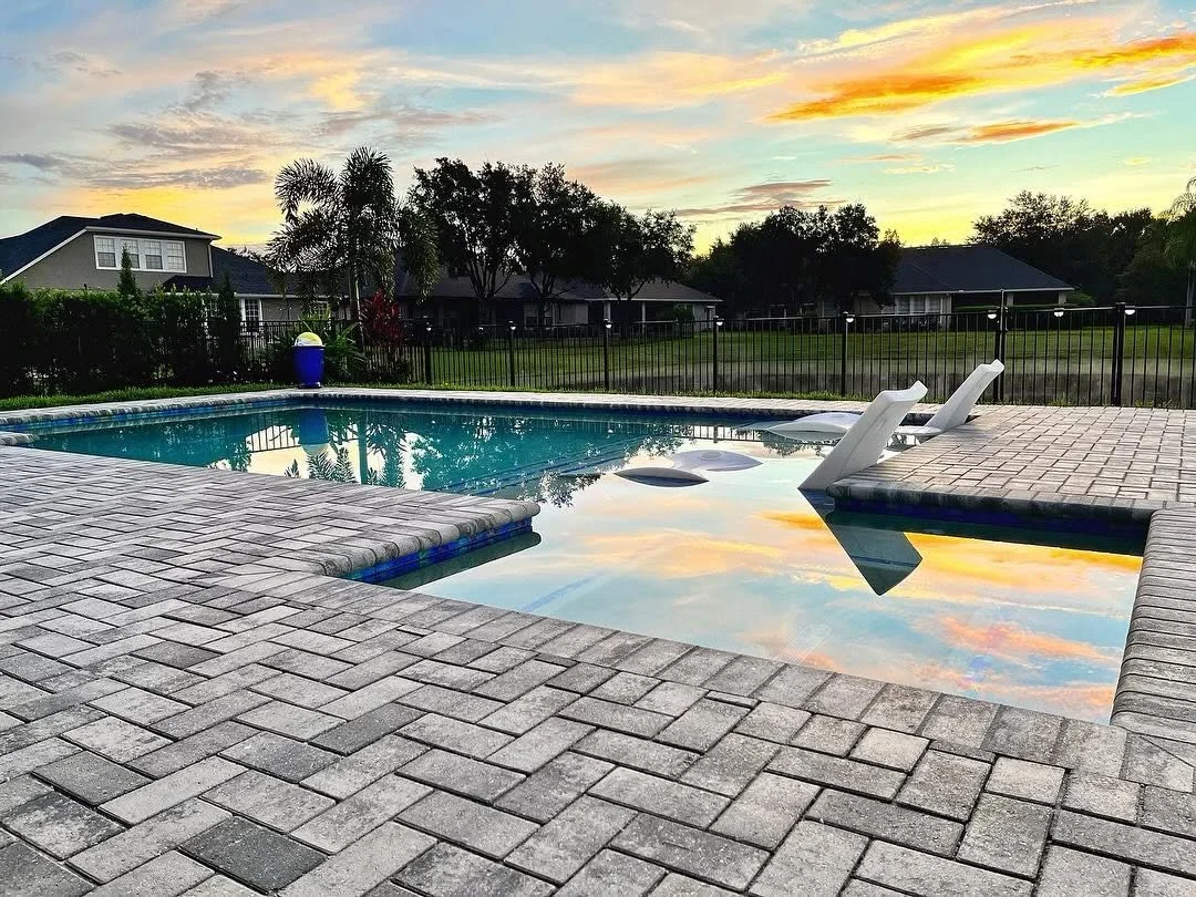 A backyard swimming pool with clear water reflecting the colorful sunset sky, surrounded by a paved stone deck, two white lounge chairs, and a landscaped background with trees and houses.