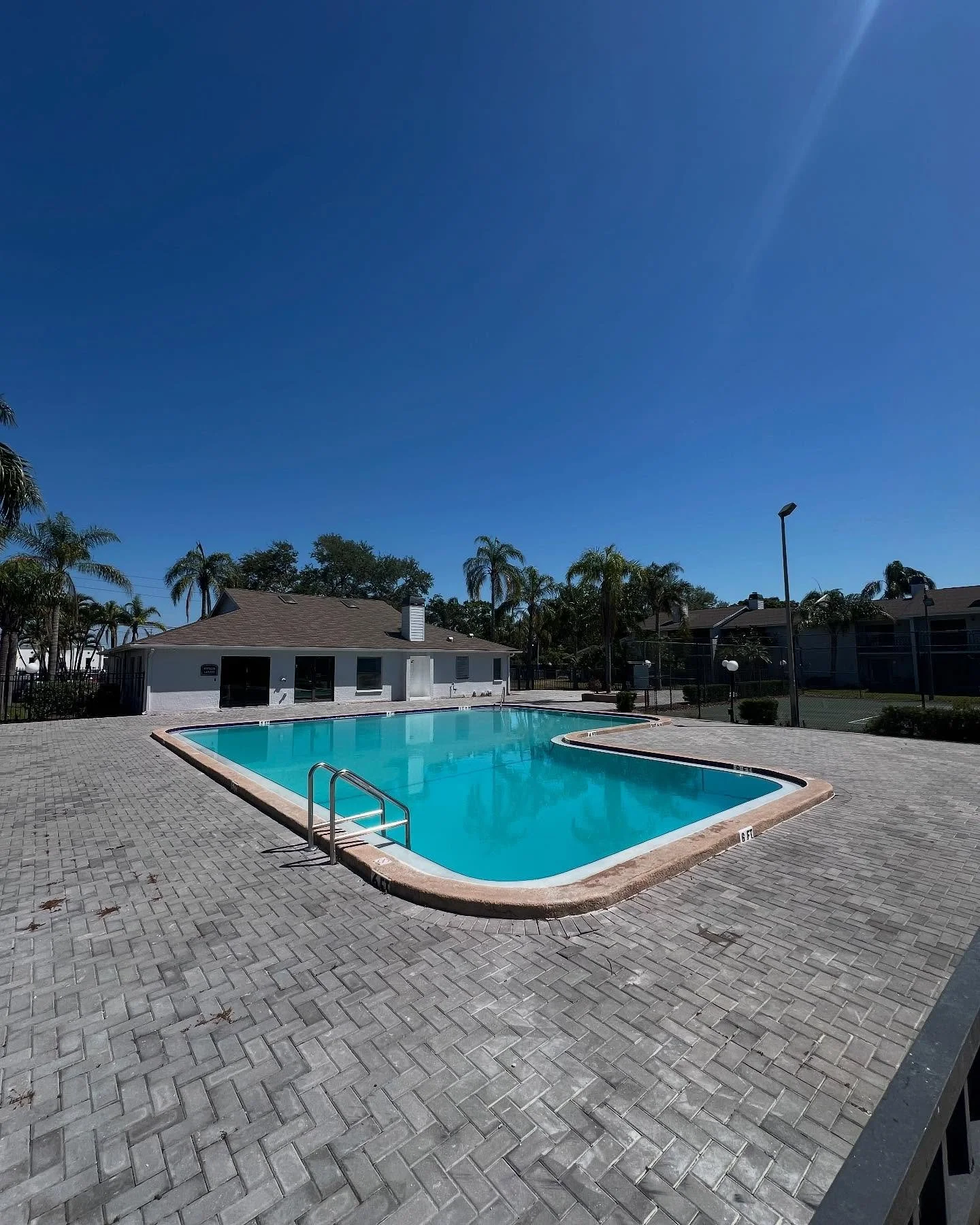 Empty outdoor swimming pool surrounded by a paved area, with a small building, palm trees, and a tall street light in the background under a clear blue sky.