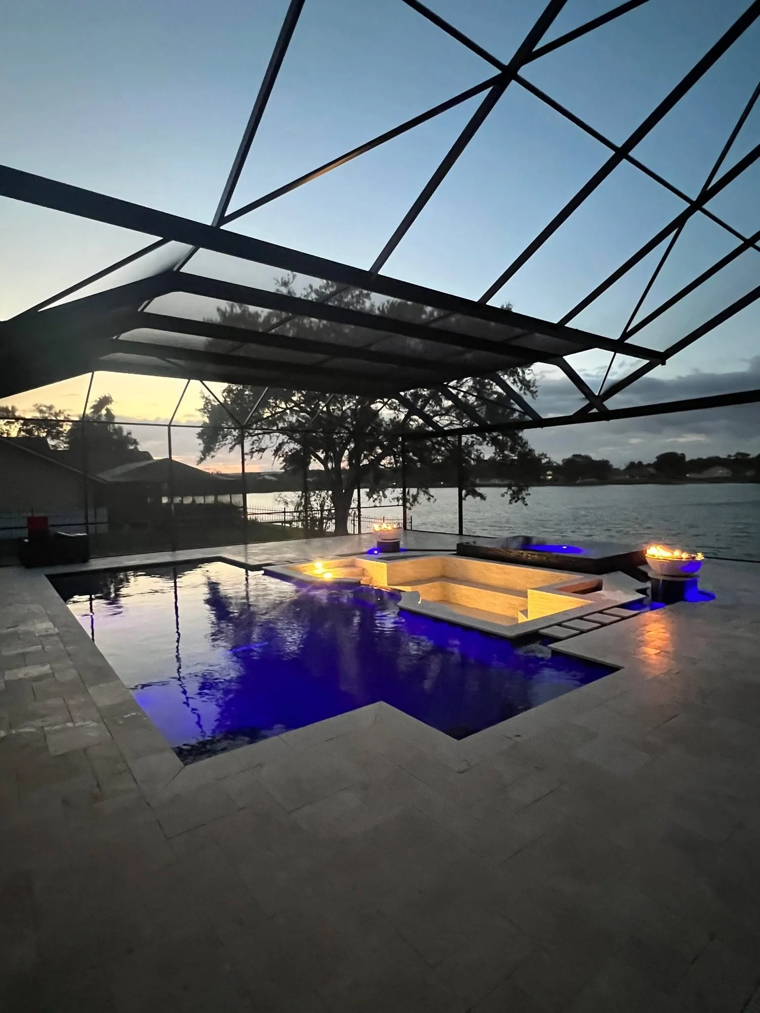An outdoor swimming pool with blue lighting at dusk, covered patio area with fire bowls, overlooking a river with trees and houses in the background.