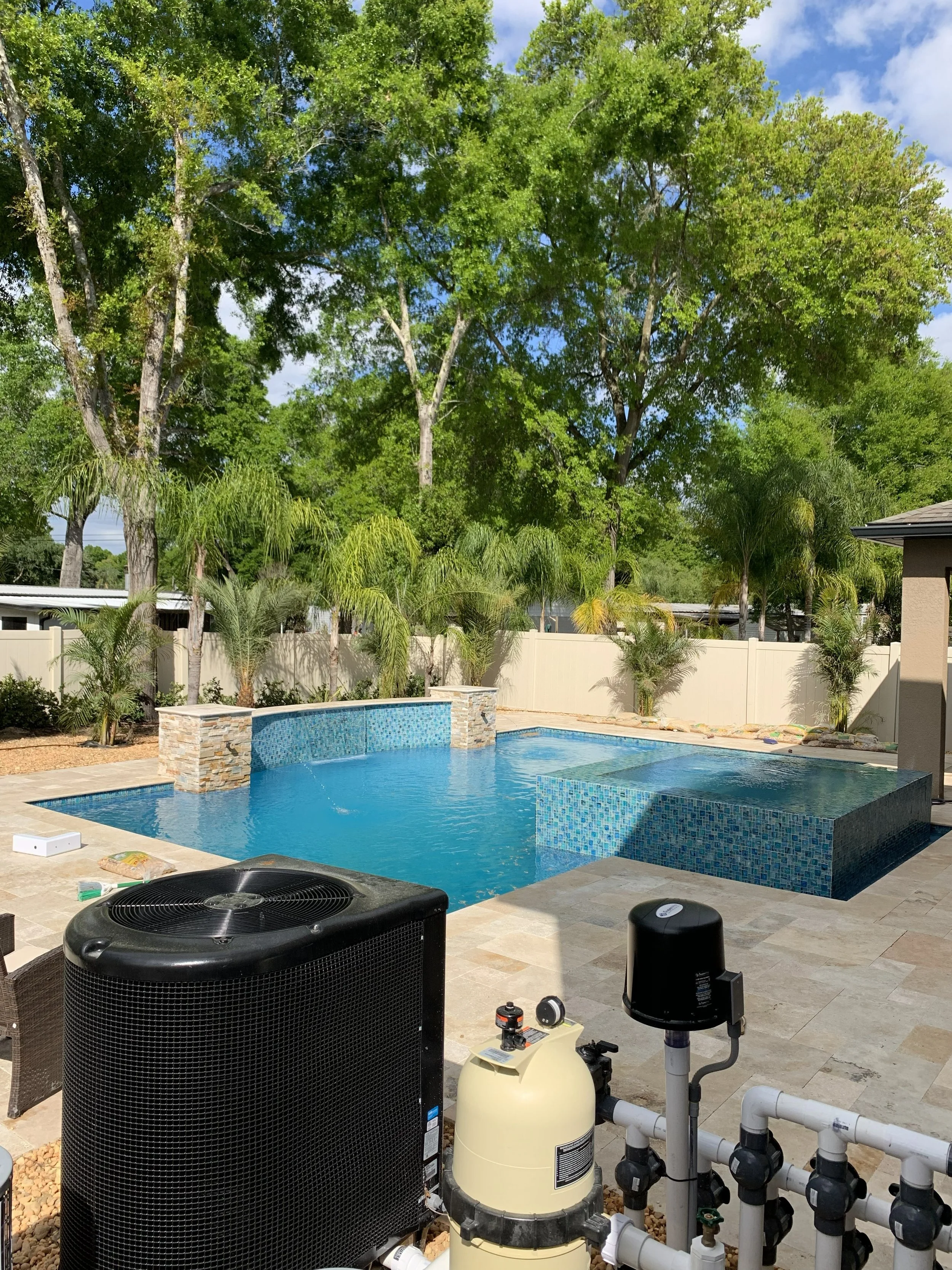Backyard pool with equipment and a fence, surrounded by tall green trees and plants.