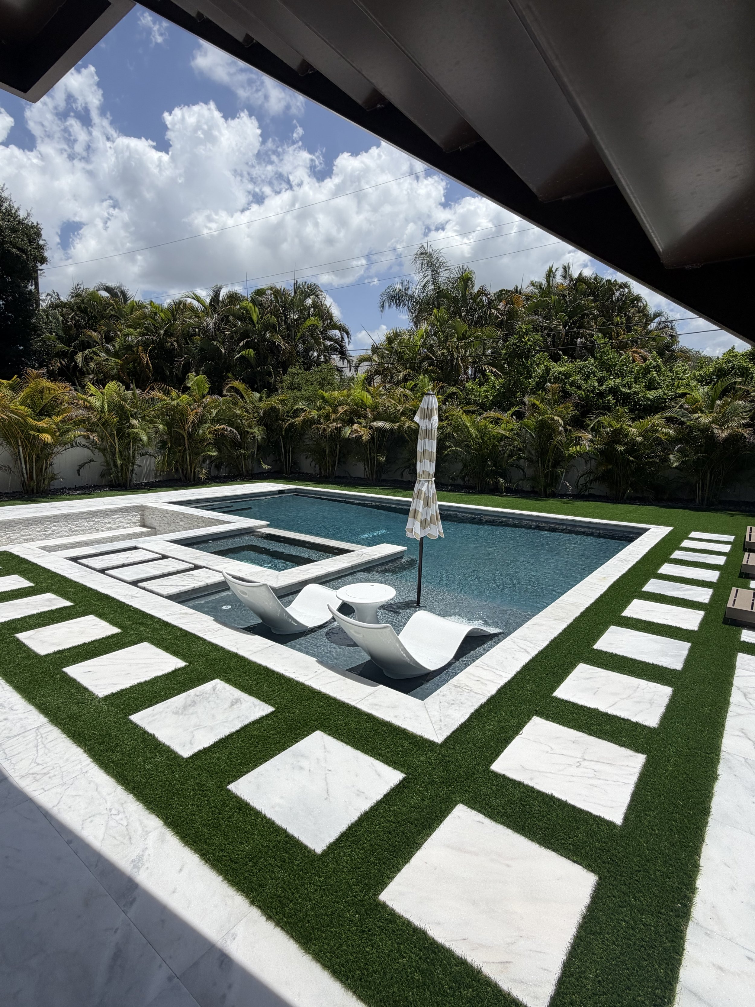 Backyard with a swimming pool, surrounded by green grass and decorative paving stones, with a tall fence and lush trees in the background, under a partly cloudy sky.