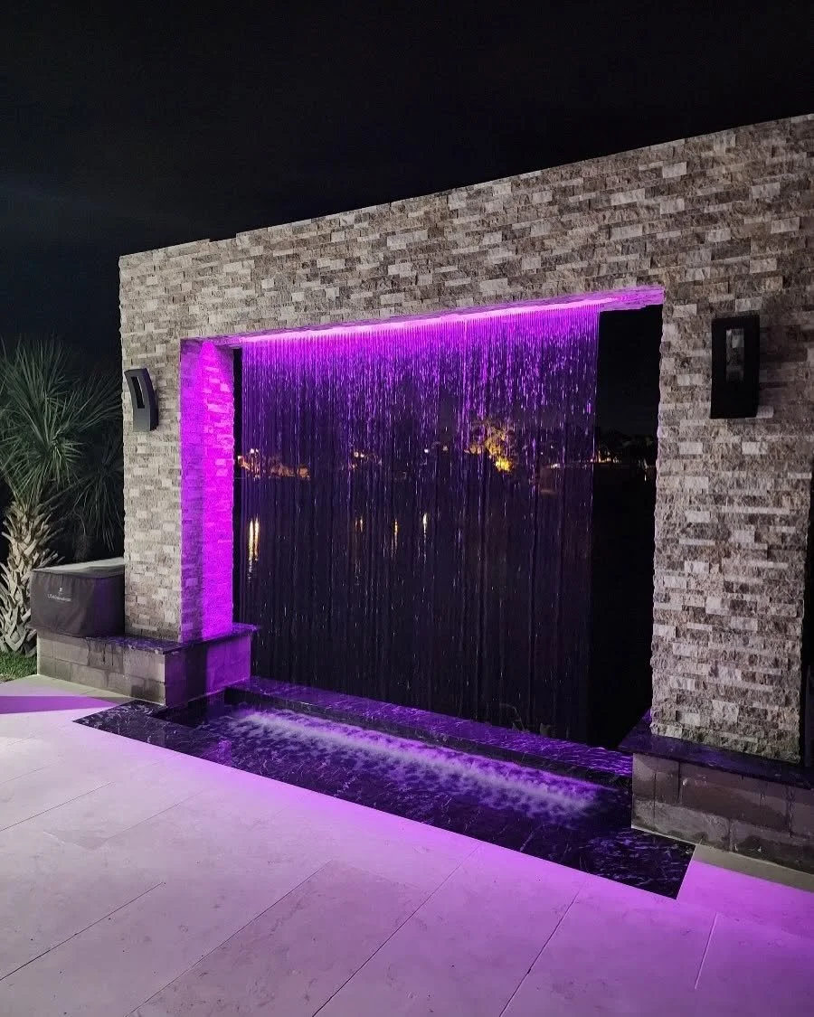 Nighttime view of an outdoor water feature with a brick wall, purple lighting, and a waterfall cascading into a shallow pool.