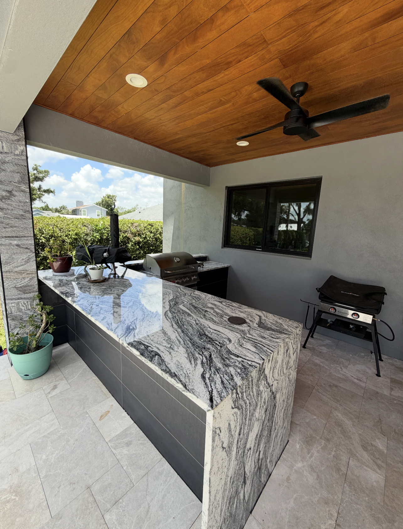 Outdoor kitchen with granite countertop, potted plants, a gas grill, and a ceiling fan, surrounded by a hedge and nearby houses visible through an open window.