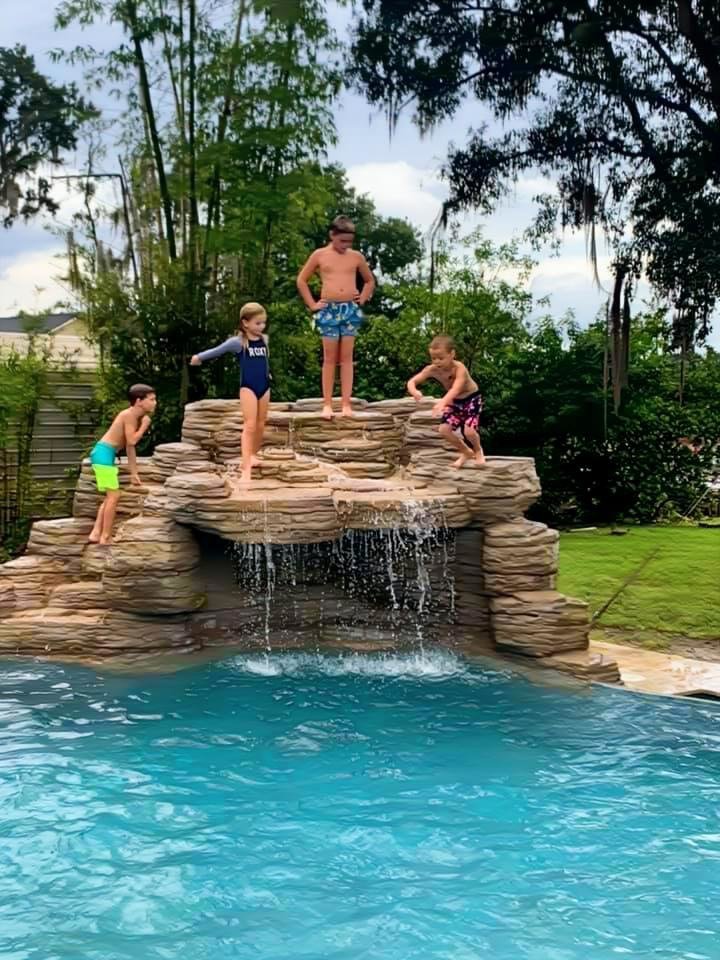 Four children playing on a rock waterfall feature above a swimming pool in a backyard, with trees and greenery in the background.