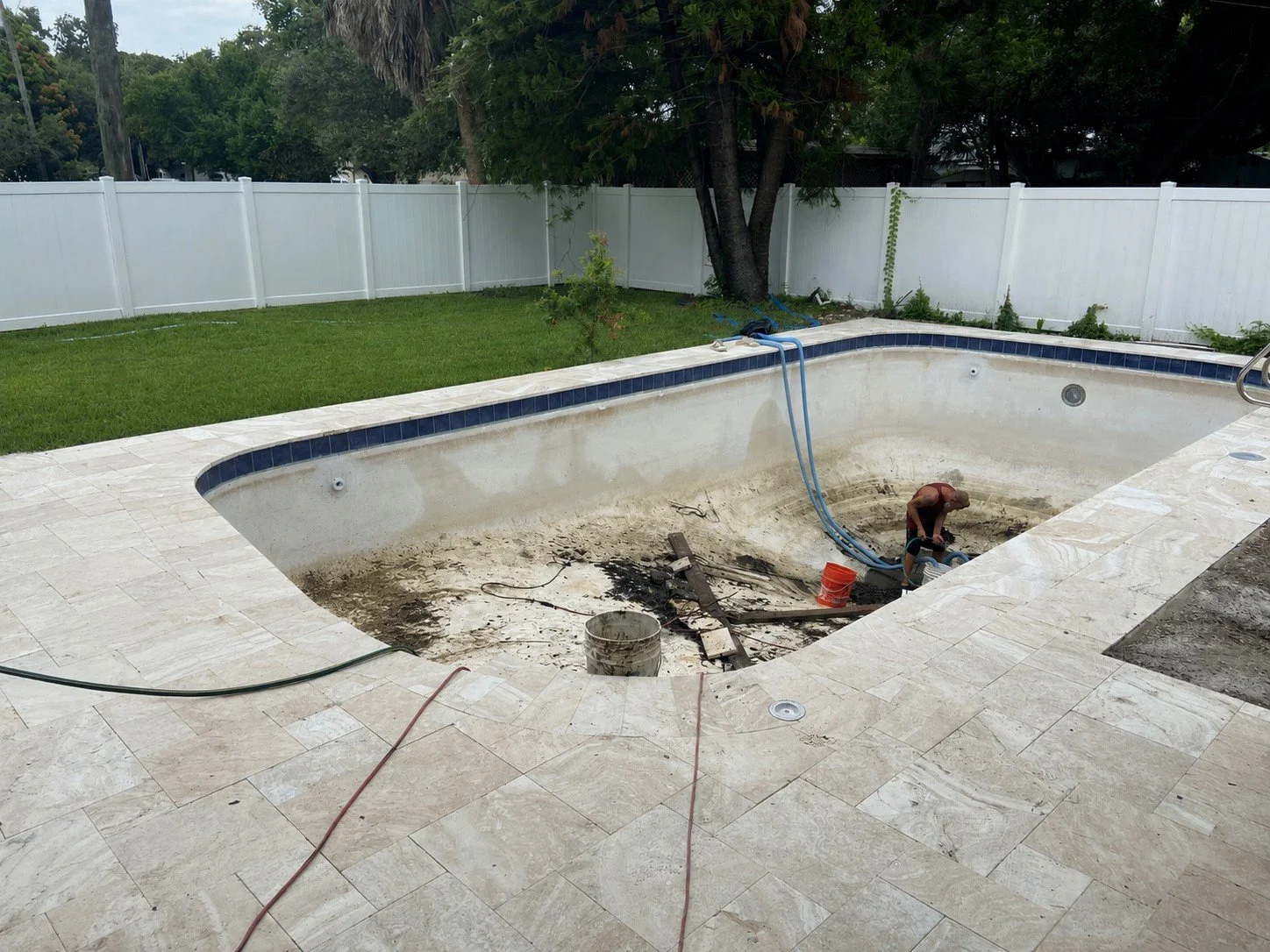 A person working in an empty swimming pool undergoing repairs, with tools, pipes, and dirt inside the pool, surrounded by a stone tiled deck and a white fence in the background.