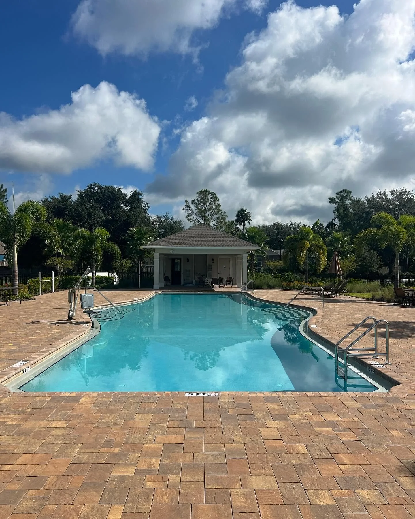 Empty outdoor swimming pool with a patio, surrounded by palm trees and lounge chairs, under a partly cloudy sky.