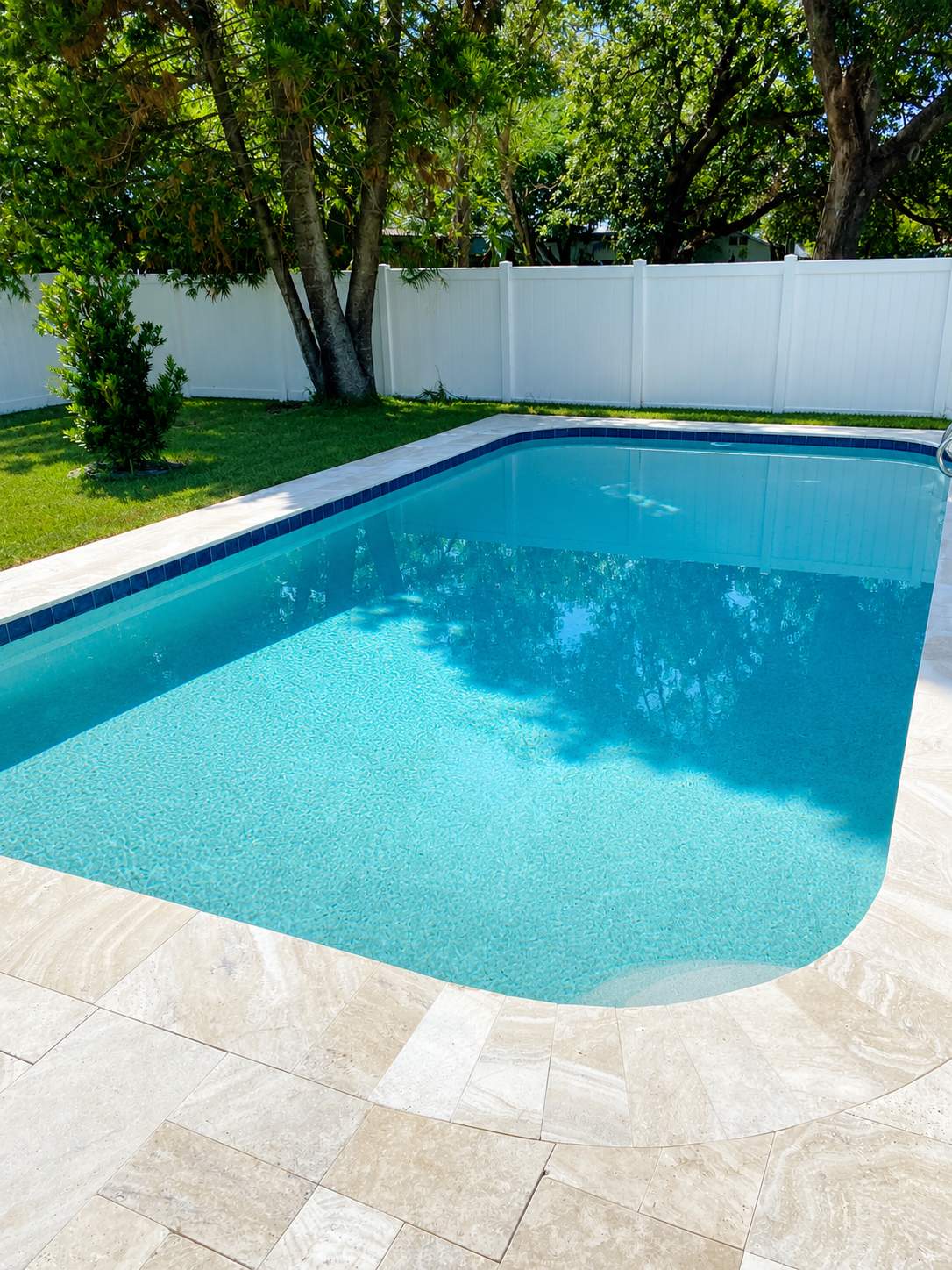 Backyard swimming pool with white tiled edge, surrounded by green grass and trees with a white fence.