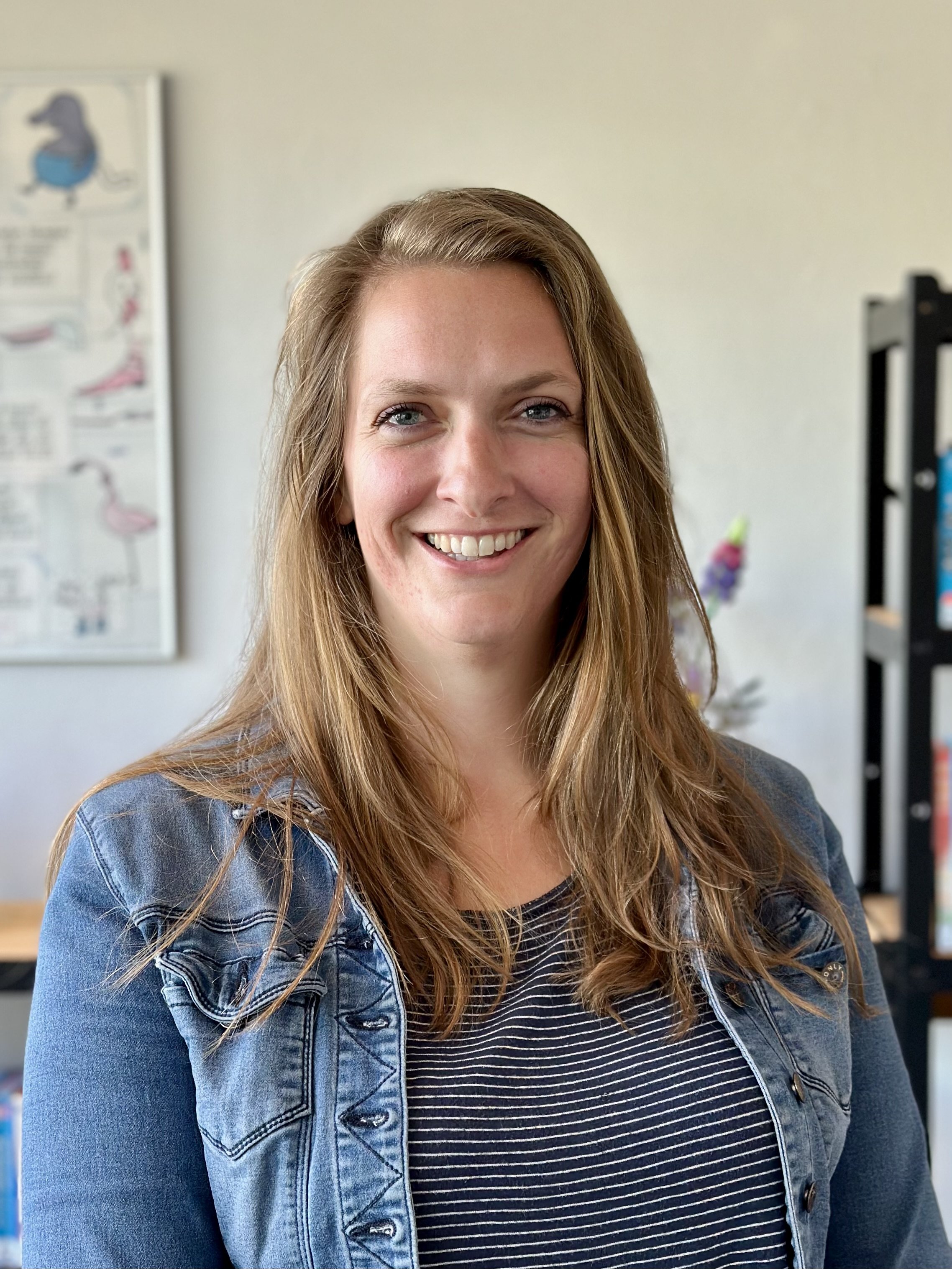 Vrouw met lang haar en een spijkerjack, binnenshuis, glimlacht. Op de achtergrond is een schilderij en een boekenplank zichtbaar.