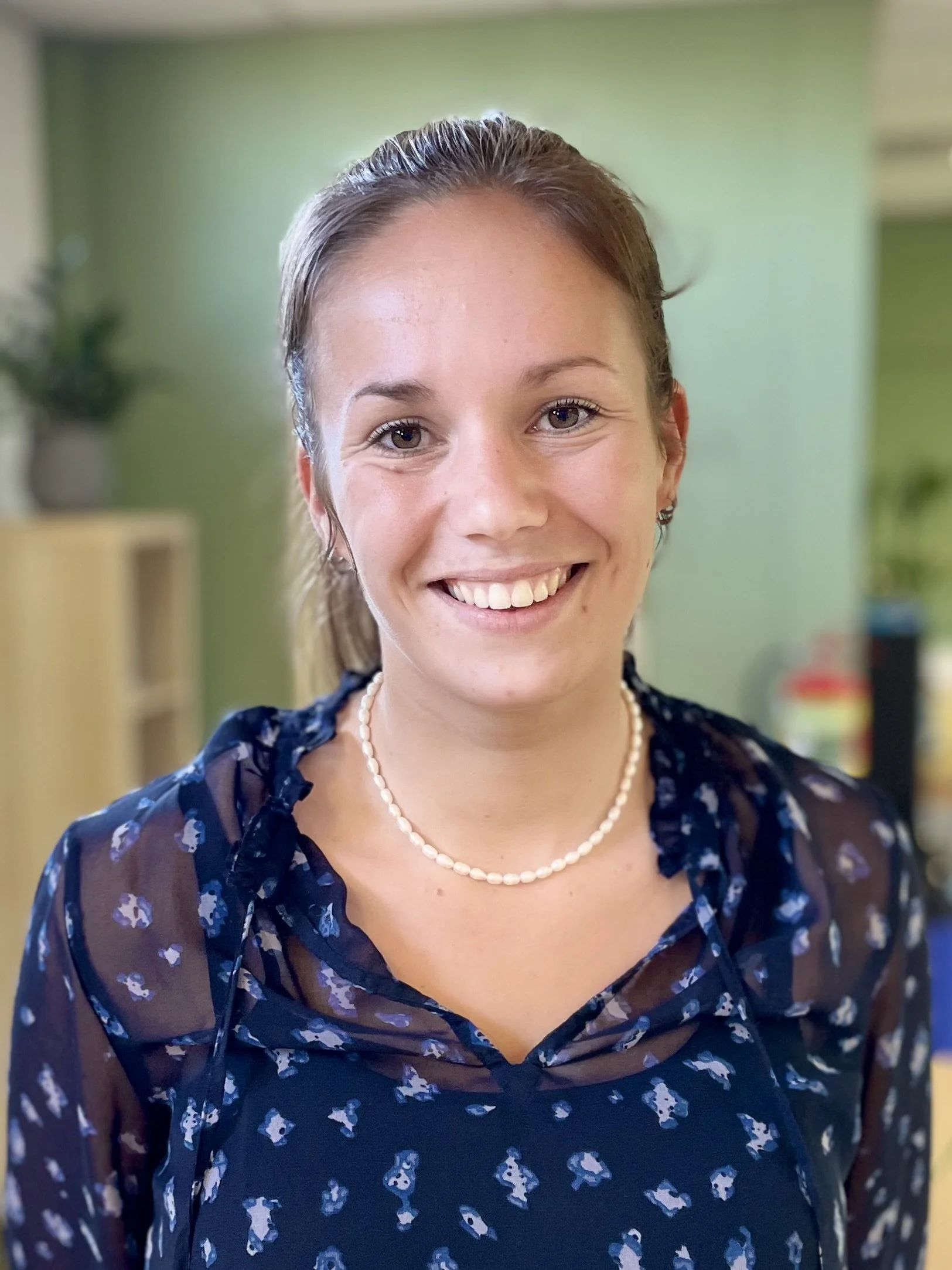 Vrouw glimlacht in kantooromgeving, draagt een blauwe blouse met patroon en een parelketting.