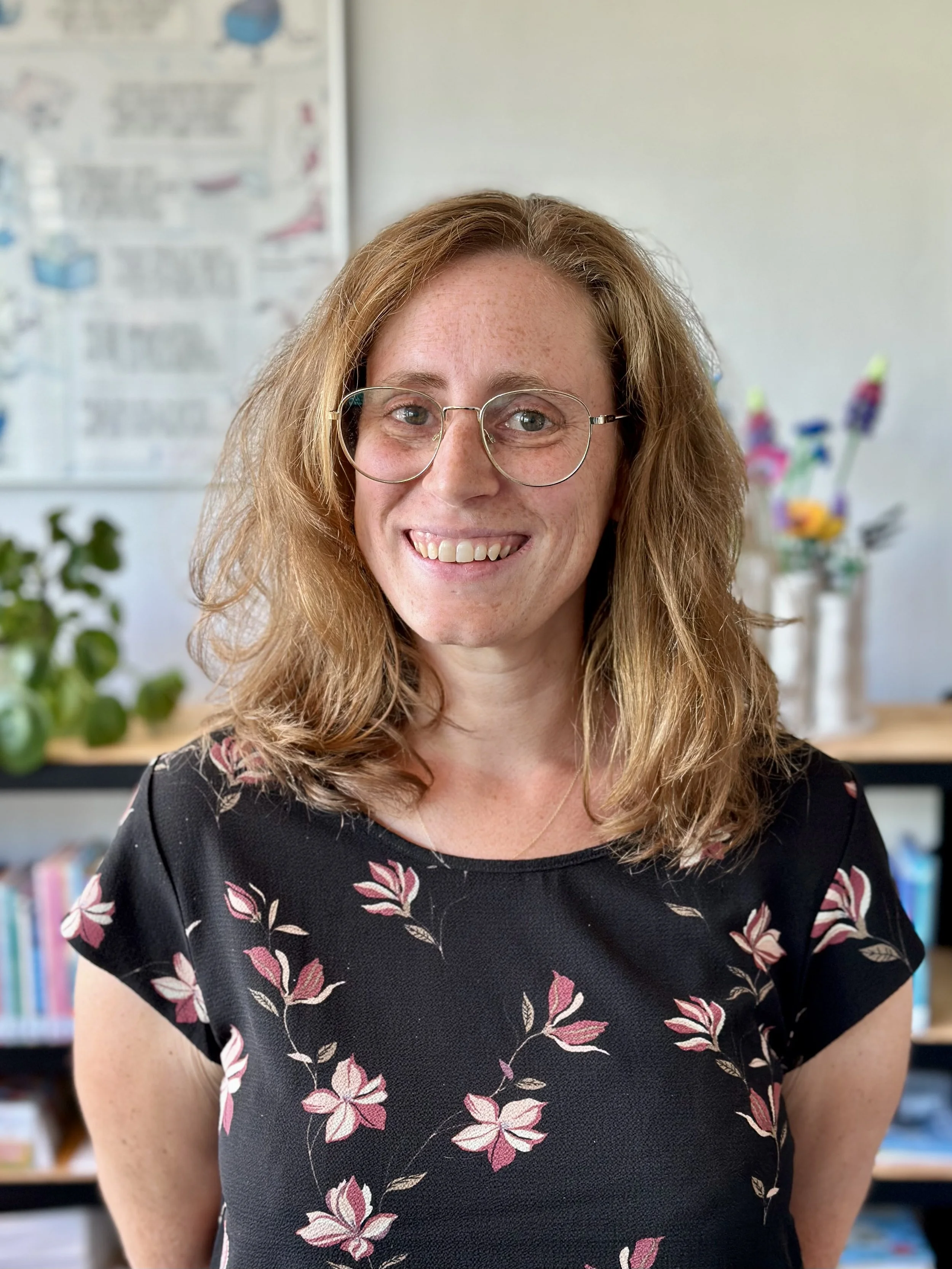 Portret van een glimlachende vrouw met een bril en halflang haar, gekleed in een zwarte bloemenjurk. Op de achtergrond zijn boekenplanken en decoratieve items te zien.