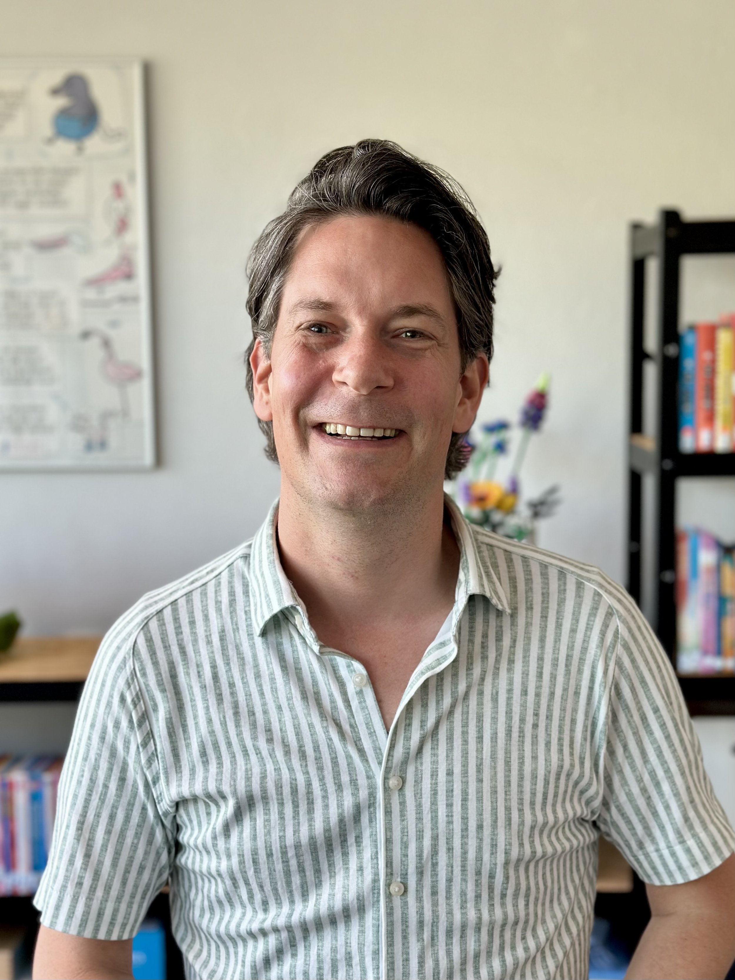 Hans de Bakker in een gestreept hemd glimlacht in een kamer in Leiden met boekenplanken en een schilderij aan de muur.