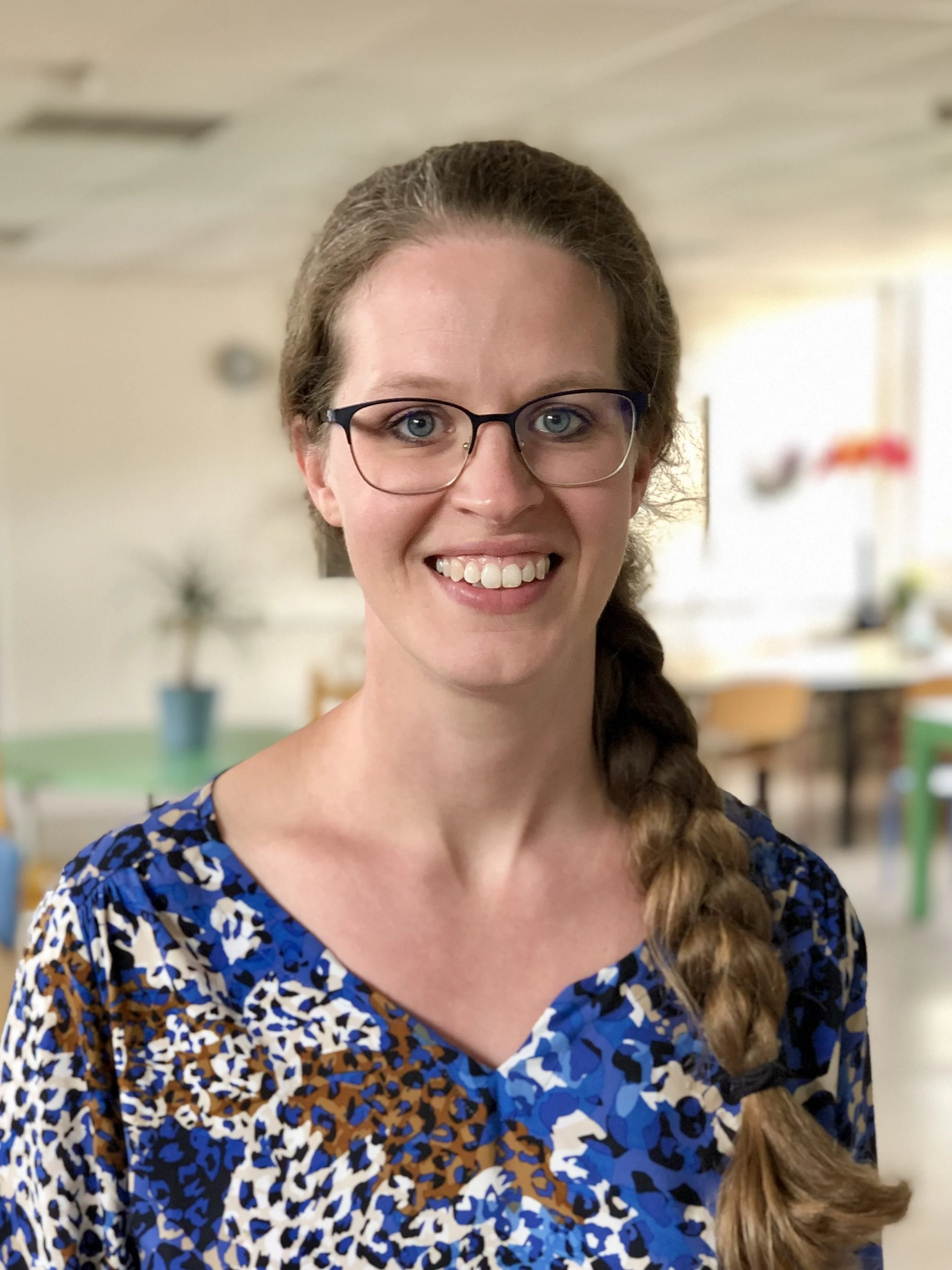 Vrouw met bril en gevlochten haar in een kleurrijke blouse lacht in een binnenomgeving.