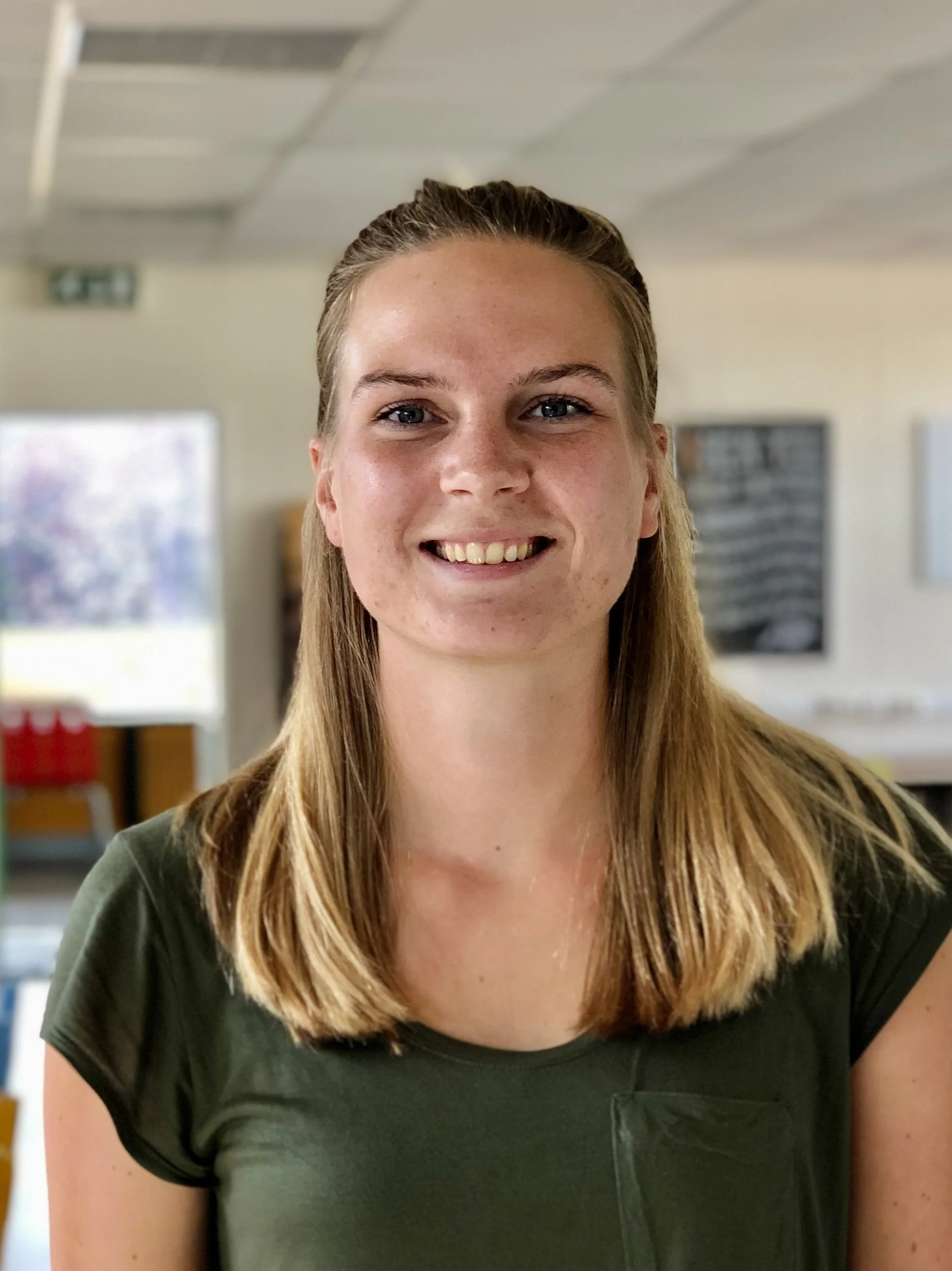 Portret van een vrouw met een groene shirt, glimlachend en staand in een binnenruimte met vervaagde achtergrond.