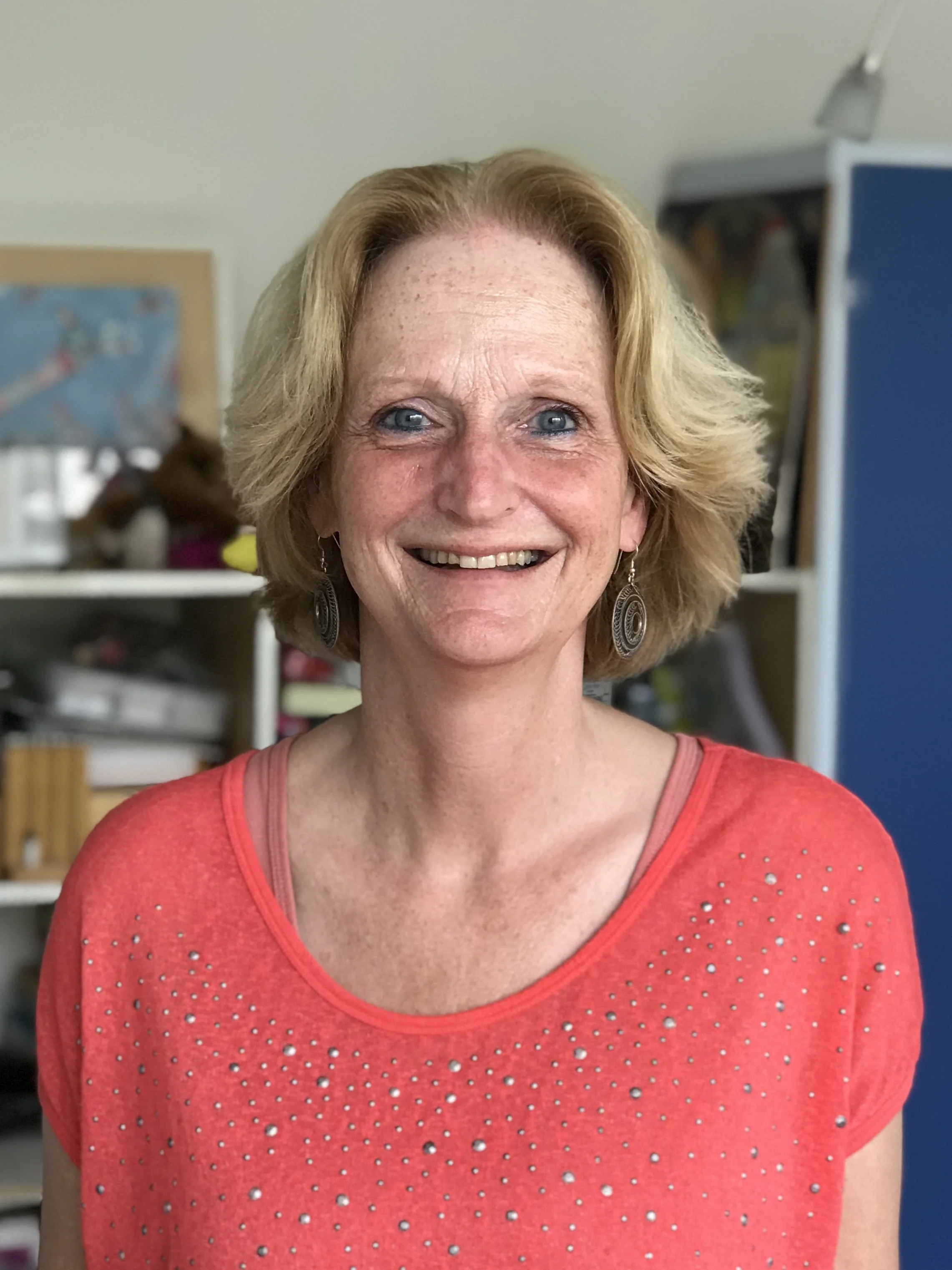 Vrouw met blond haar en rode trui glimlacht in een kamer.