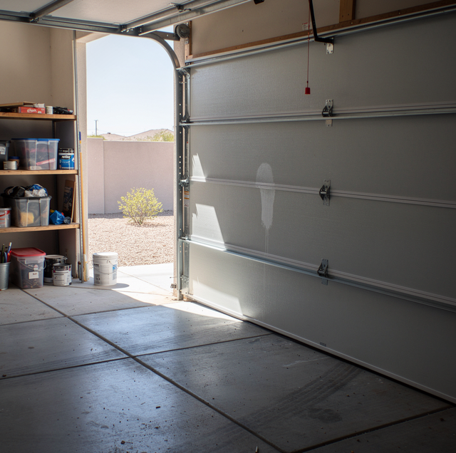Open garage door reveals concrete floor, wooden shelves with storage containers and paint cans, and a view of desert landscap