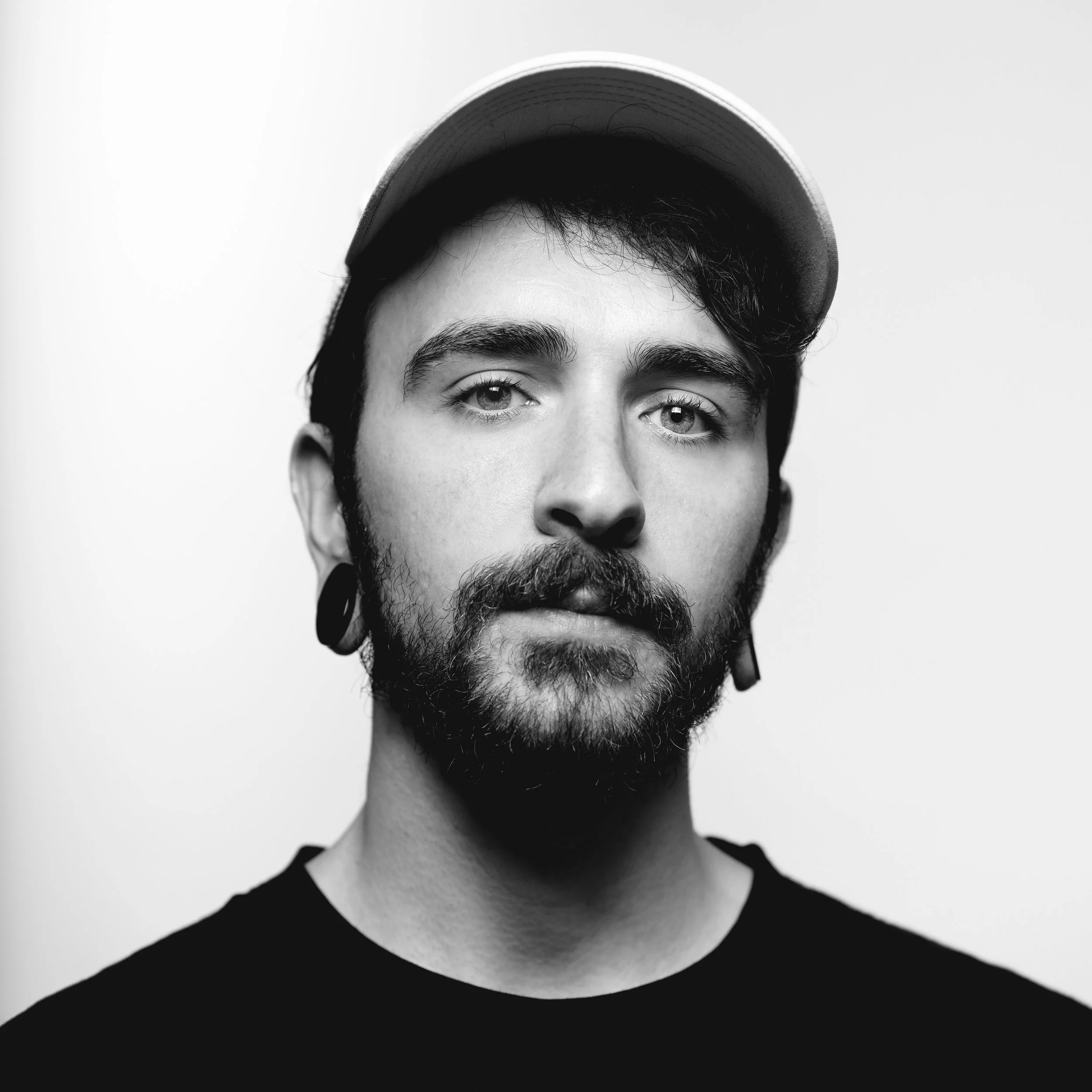 A black and white portrait photo of a young man with a beard and mustache, wearing a cap and large earrings, looking directly at the camera with a neutral expression.