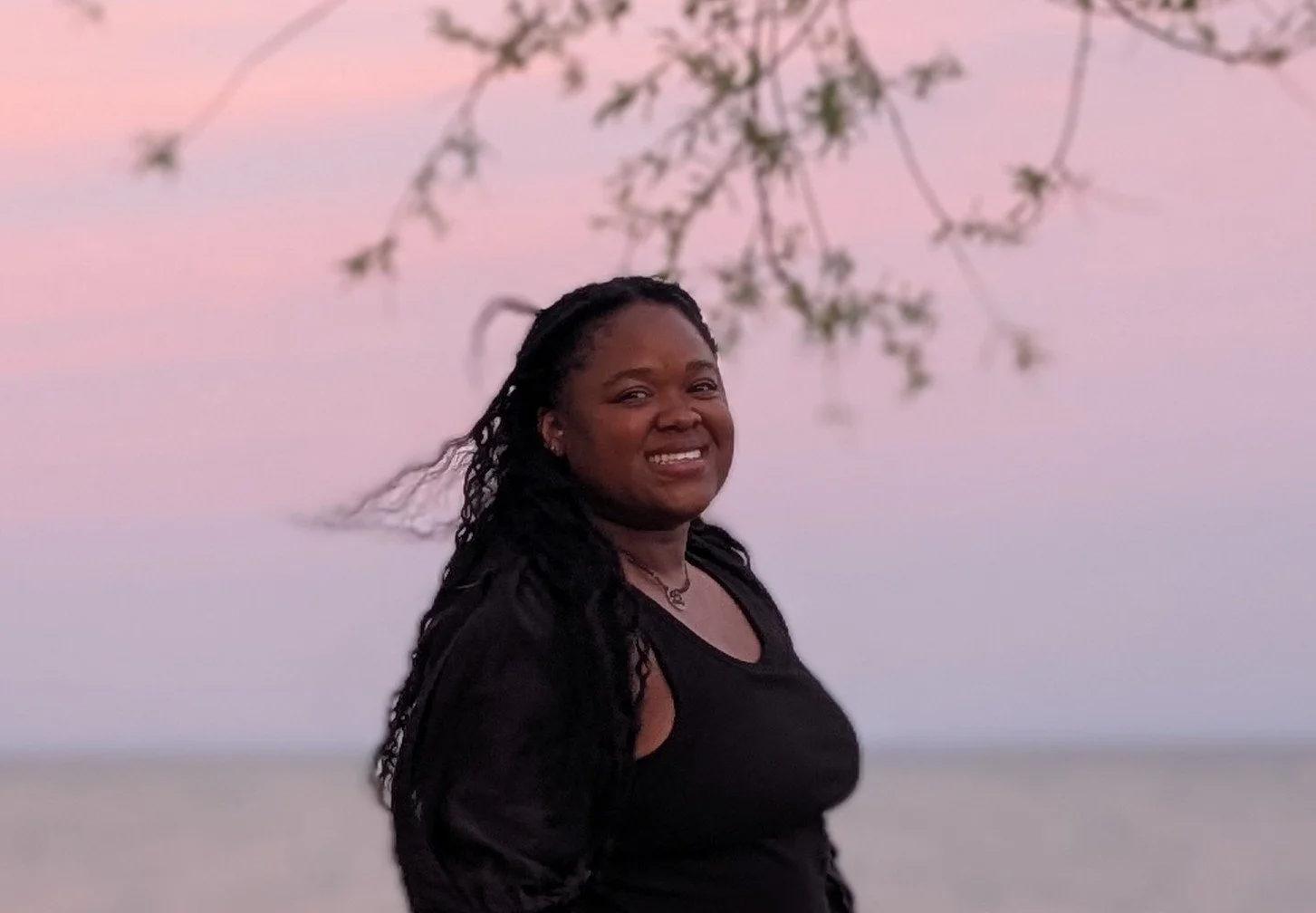 A smiling woman with long curly hair standing outdoors during sunset, with pink and purple sky and bare tree branches in the background.
